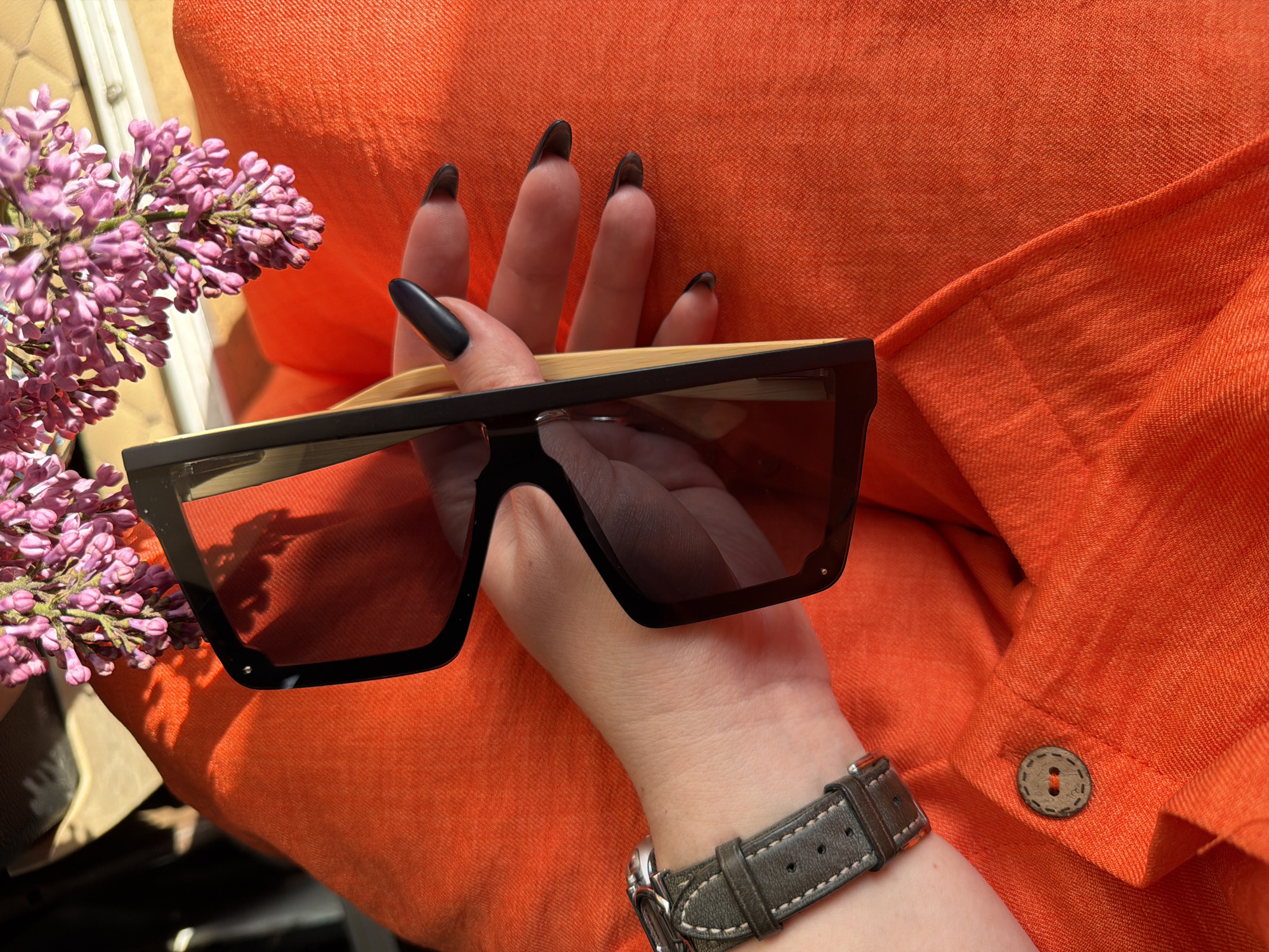 Here's a detailed description of the image:
**Subjects:**
* A woman's hand is holding a pair of sunglasses. The nails are long and painted black.
* A wristwatch with a gray leather strap is visible on the wrist.
**Objects:**
* A pair of sunglasses with a black frame and dark lenses.
* A branch of lilac flowers is positioned to the left of the hand.
* An orange-colored garment or fabric is in the background. A button is visible on the garment.
**Environment:**
* The location appears to be outdoors, indicated by the lighting and natural elements.
**Other Details:**
* The hand is posed to display the sunglasses.
**Location:**
* The city and specific location are unknown.