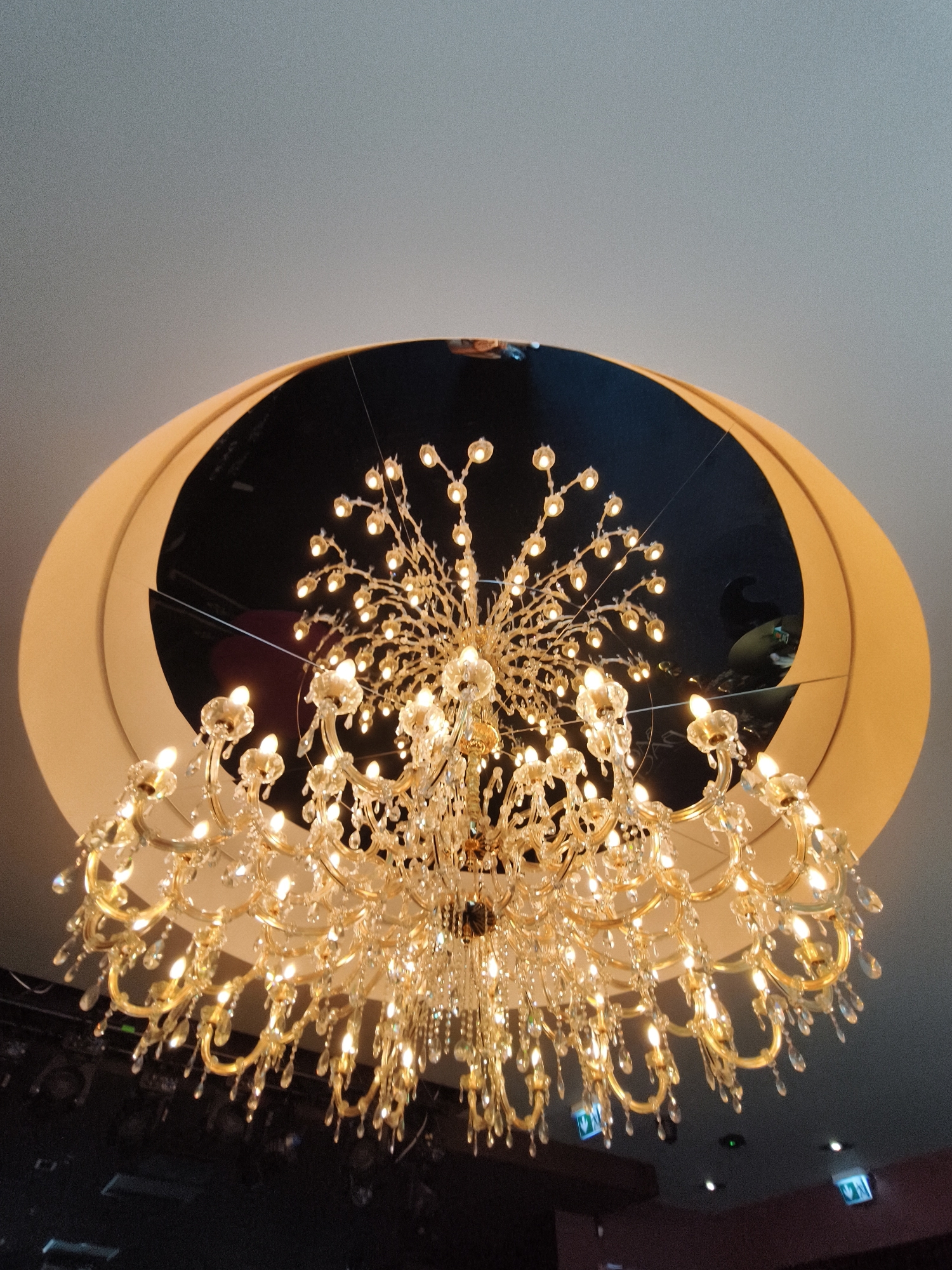 A chandelier hangs from the ceiling of a building. The chandelier is gold-colored with many crystal accents and light bulbs. It is located in Tallinn, Estonia. The ceiling has a large oval-shaped cut-out with a reflective surface inside. The overall impression is one of luxury and elegance.