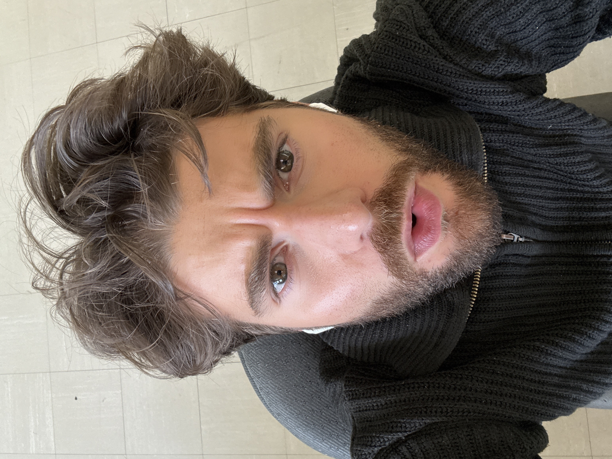 The image shows a man with brown hair and a beard, wearing a black sweater. He is looking directly at the camera. The background appears to be a tiled floor. There is no indication of the city of Montreal.