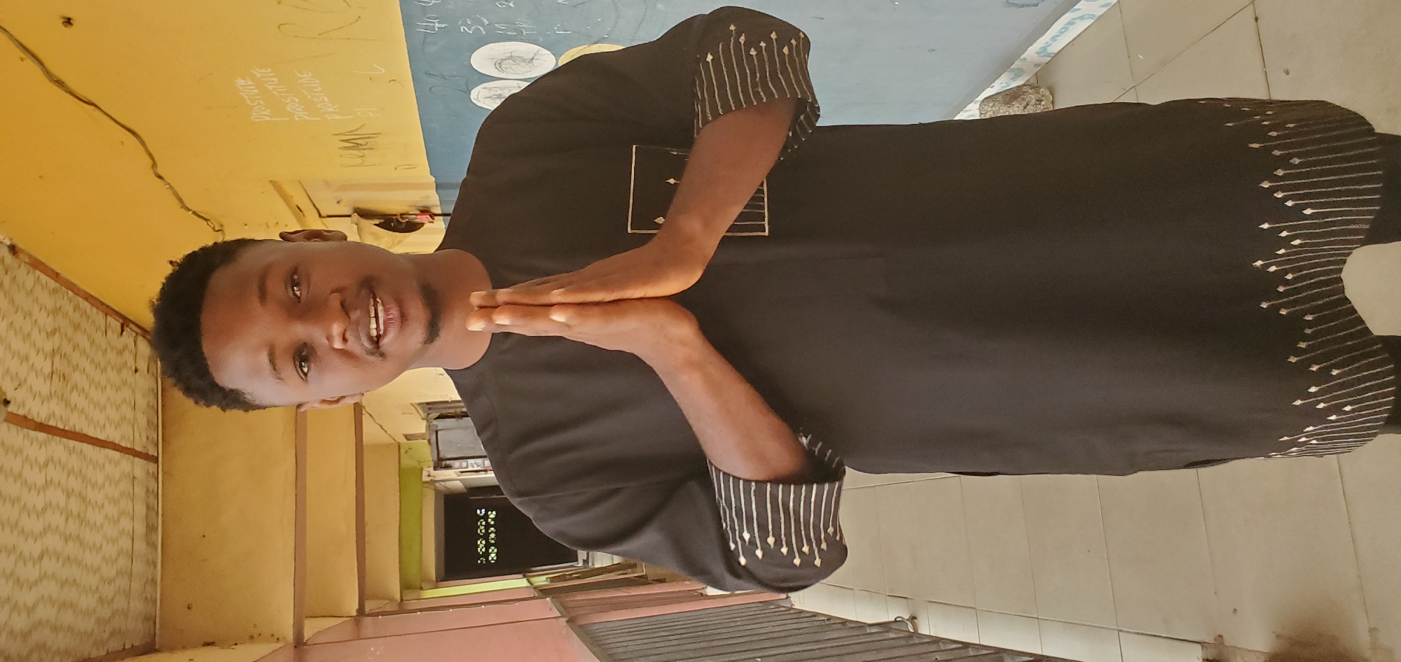 The image shows a man in Mararaba, Nigeria. He is wearing a black, long-sleeved shirt with white stripe patterns on the cuffs and a chevron pattern around the bottom hem. The man has short black hair, and he is smiling. He is holding his hands together in front of him. The background includes an exterior wall with paint or writing on it, and a tiled floor. There is what appears to be a corrugated iron roof or awning above him, and a barred opening behind him.