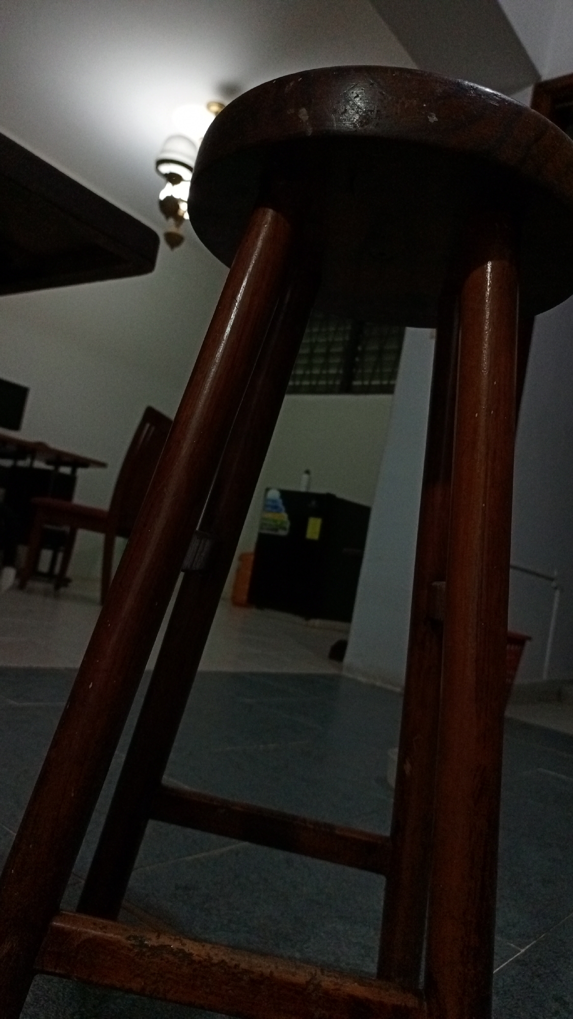 The image depicts a close-up view of a dark wooden stool with three legs. The room is dimly lit, with a dining table and chairs visible in the background. There is also a refrigerator and other household objects present in the back. The location in the city of Los Robles, Venezuela cannot be determined from this image.