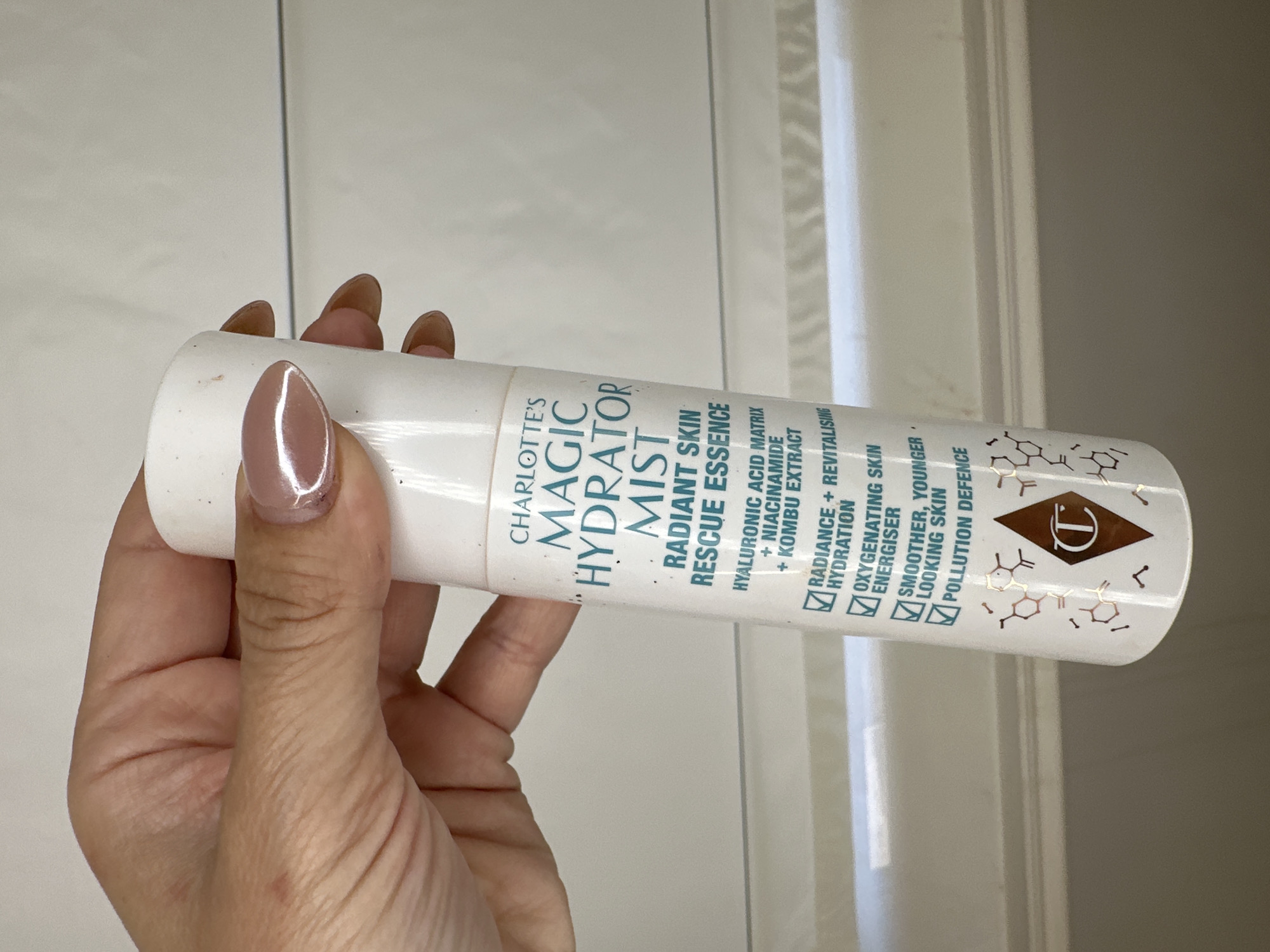 A left hand with light-toned skin and manicured nails, painted a glossy nude-brown, securely grips a white cylindrical bottle. The bottle's label, featuring teal text, identifies the product as 'CHARLOTTE'S MAGIC HYDRATOR MIST RADIANT SKIN RESCUE ESSENCE,' noting key ingredients like 'HYALURONIC ACID MATRIX + NIACINAMIDE + KOMBU EXTRACT.' Four checked bullet points list product benefits: 'RADIANCE + REVITALISING HYDRATION,' 'OXYGENATING SKIN ENERGISER,' 'SMOOTHER, YOUNGER LOOKING SKIN,' and 'POLLUTION DEFENCE.' Towards the base, a copper-toned diamond logo with a stylized 'C' is visible, surrounded by scattered molecular structure motifs in the same metallic finish. The background is a light, possibly off-white or light gray surface, with distinct horizontal lines suggesting tiling or a textured wall, creating an indoor, well-lit environment.