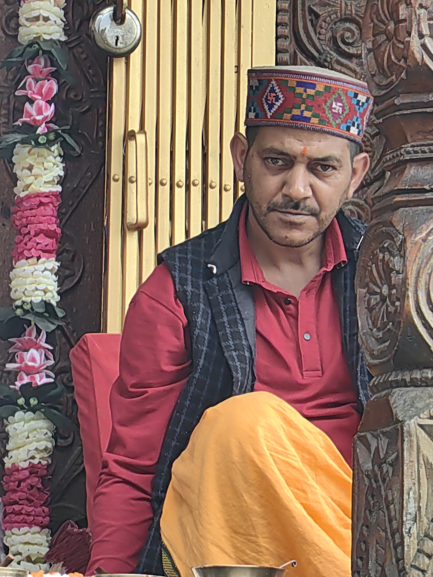 In Manali, India, a man with a mustache, a red tilak on his forehead, and a patterned cap sits in front of a golden accordion gate and carved wooden structure. He wears a red polo shirt, a dark checkered vest, and an orange cloth draped over his lap. To his left, a garland of pink and white flowers hangs. The scene is illuminated by natural light, creating shadows on the man's face and the surrounding objects.