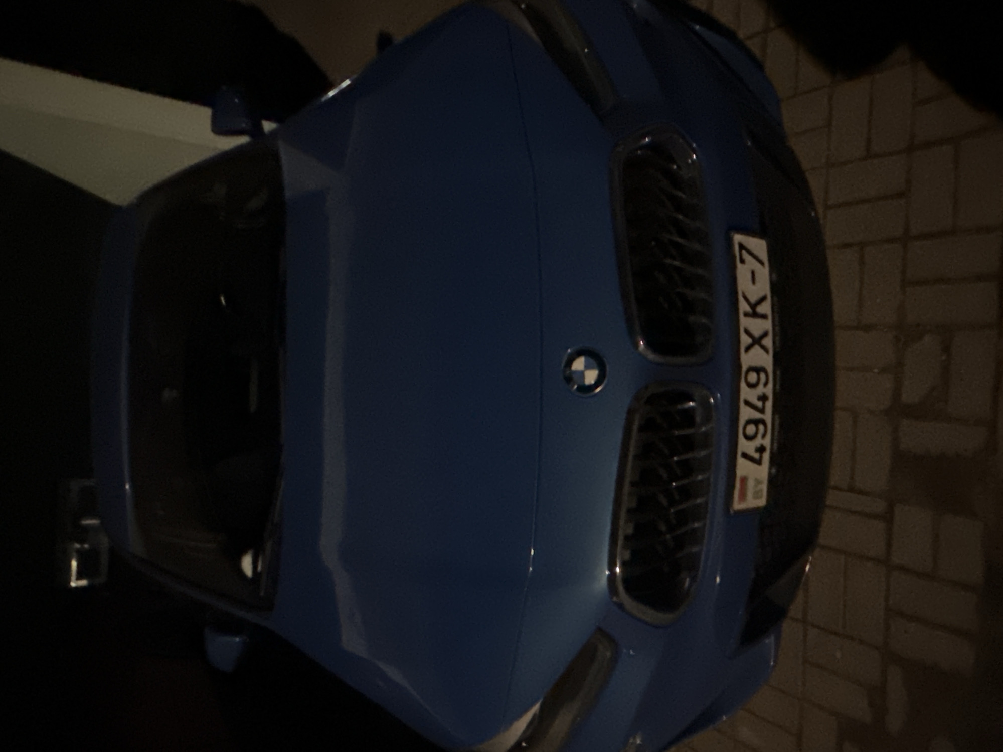 The image shows a blue BMW parked on a brick-paved surface, likely a sidewalk or driveway. The car's license plate, "4949 XK-7," is visible, indicating it is registered in Belarus. Based on the license plate, the car is likely located in Minsk, Belarus. The car door is open. The scene is illuminated by artificial lighting, suggesting it is nighttime or low-light conditions.