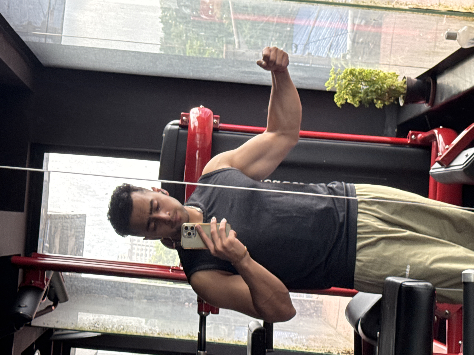 The image shows a man in a gym, taking a mirror selfie. He is wearing a sleeveless black shirt and light brown sweatpants. He is holding a smartphone in his left hand, and his right arm is flexed, showing his bicep. In the background, there is gym equipment, a window with a view of buildings, and a small potted plant. The gym appears to be modern and well-equipped. The man's attire and the gym environment suggest he is engaged in a workout routine. There is no indication of the specific location within Kolkata, India.