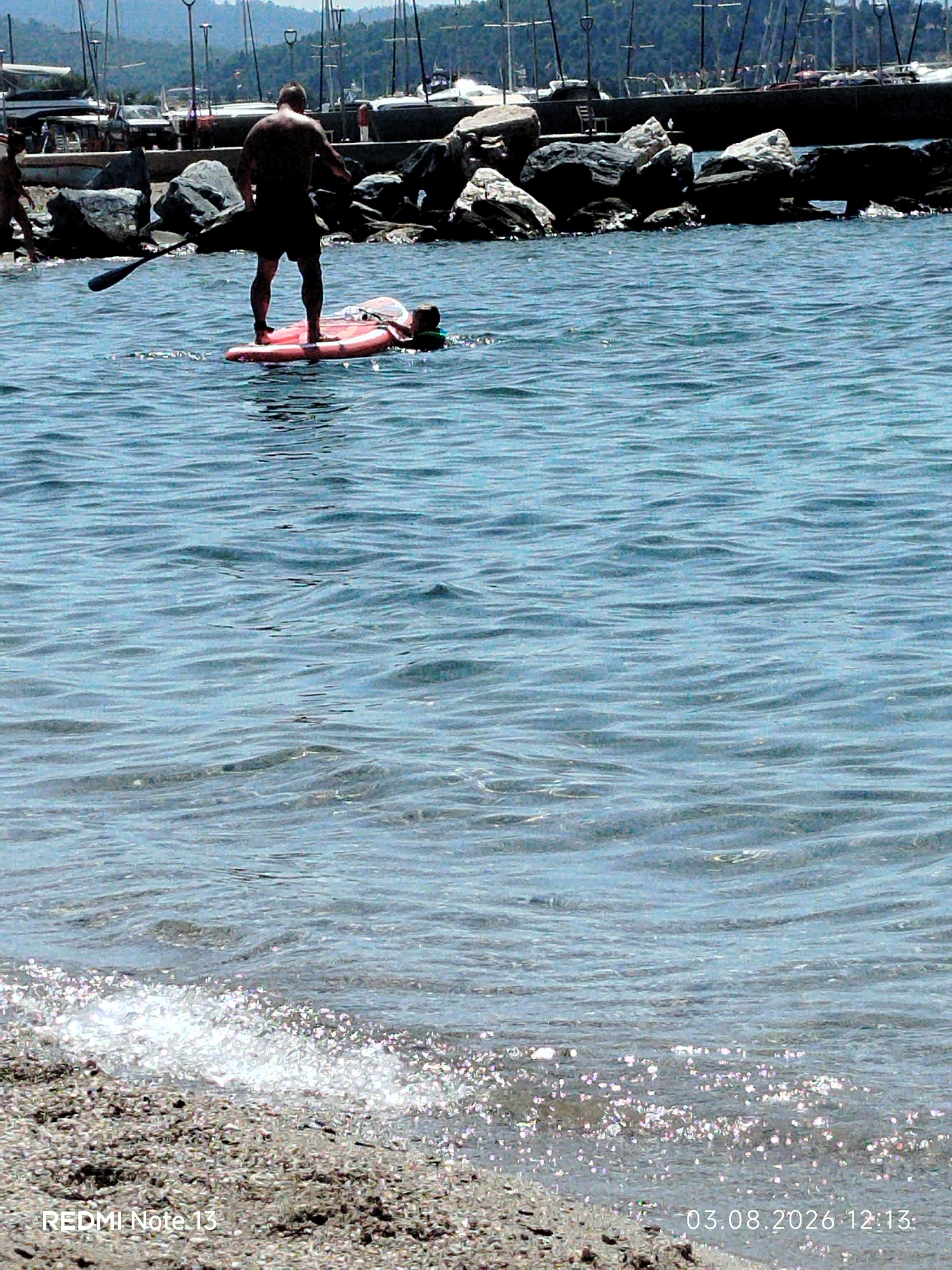 In Sithonia Municipal Unit, Greece, a man stands on a pink paddleboard in the water, holding a paddle. He is wearing dark shorts. Another person, partially visible, is lying on the paddleboard behind him. The background shows a harbor with docked boats and a rocky shoreline. The water is blue and appears slightly choppy. A portion of a beach with pebbles and small shells is visible in the foreground, with the edge of the water lapping onto it. A timestamp in the lower right corner indicates "03.08.2026 12:13".