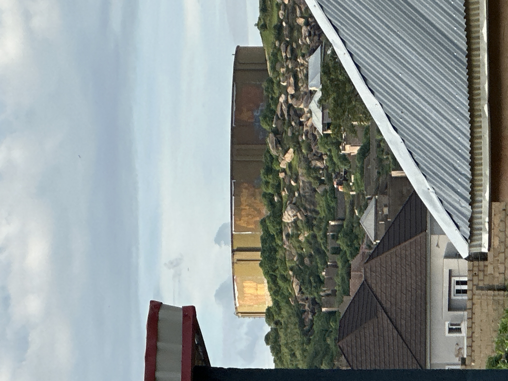 The image shows a large, circular water tank perched atop a rocky, tree-covered hill. The tank is a weathered yellow and orange color, with visible vertical seams. Below the hill, several houses with dark, pitched roofs are visible, along with a brick wall in the foreground. The sky is cloudy, with patches of blue peeking through. A small, dark object, possibly a bird or a small aircraft, is visible in the sky above the water tank. The scene appears to be an outdoor landscape in the city of Jos, Nigeria, characterized by its hilly terrain and residential architecture. The image captures a static moment with no apparent human activity or interaction.