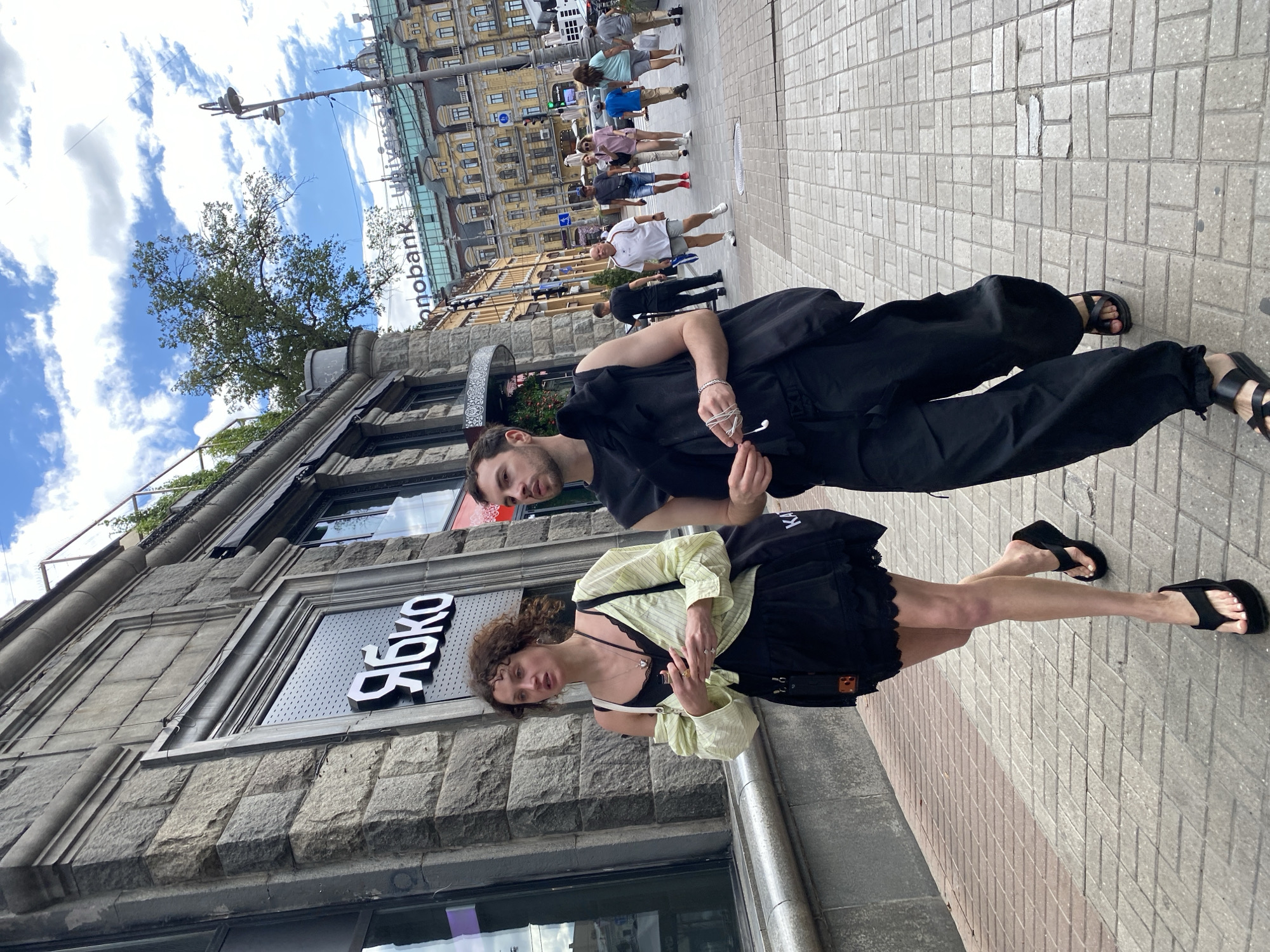 In an urban environment, two individuals, a woman and a man, walk side-by-side on a pedestrian sidewalk. The woman, on the left, has curly brown hair and is wearing a black dress with lace trim, a light yellow open-buttoned shirt, and black platform flip-flops. She carries a black tote bag over her shoulder. The man, on the right, has short brown hair and is dressed in a black sleeveless vest over a black sleeveless top, black wide-leg pants, and black slide sandals. He holds white earphones in his right hand. They are walking towards the right side of the frame.
Behind them, other pedestrians are visible on the sidewalk, and buildings line the street. One building has a sign with the Cyrillic letters "ЯБКО" in white. Further in the background, a modern building with the word "monobank" and "Universal" is visible. The sky is blue with scattered white clouds. The scene is captured from a slightly low angle.