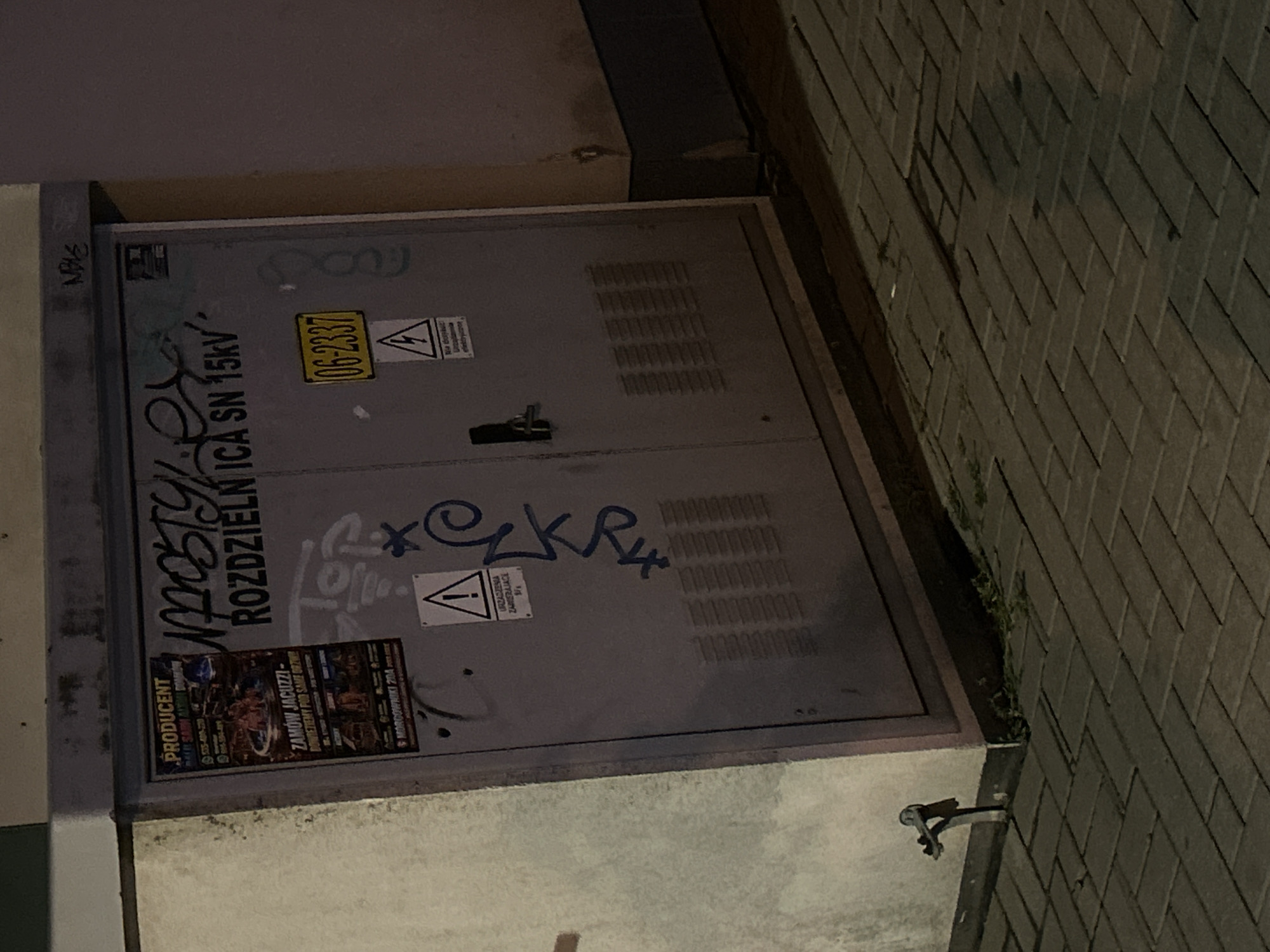 The image shows a utility box on a sidewalk in the city of Siedlce, Poland. The utility box is gray and appears to be made of metal. It is covered in graffiti and has several stickers and labels on it. One sticker is an advertisement for "PRODUCENT" offering "ZAMÓW JACUZZI - DOWIEZIEMY POD SAME DRZWI". Another sticker is a yellow rectangle with the number "06-2337" printed on it. Above that, there is a warning sign with a lightning bolt symbol. The utility box is located next to a building with a rough textured wall. The sidewalk is paved with brick-like stones. In the foreground, there is a metal pipe or faucet attached to the wall. The lighting suggests it is nighttime or dusk.