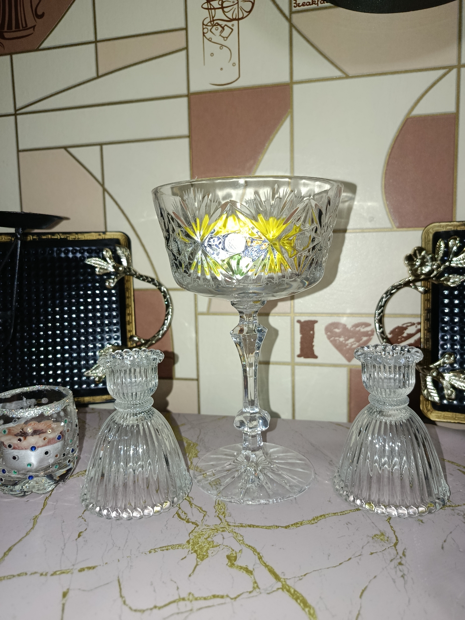 The image depicts a still life arrangement of decorative glass items on a pink marble-patterned surface. In the center, a tall, stemmed glass goblet with intricate crystal cutting is prominently displayed. To its left and right are two smaller, bell-shaped glass candle holders with fluted designs. To the far left, a small, cylindrical glass votive holder adorned with glitter and colorful beads is visible. Behind the glass objects, a patterned tile backsplash with geometric shapes in white, pink, and tan, separated by thin gold lines, forms the background. On the left side of the background, a black square object with a metallic gold decorative handle and a perforated surface is partially visible. To the right, a similar decorative object is also partially visible. The scene is well-lit, casting subtle shadows on the surface. The overall presentation suggests an indoor domestic setting, possibly a display on a table or shelf. The image does not provide any information or context related to Tashkent, Uzbekistan.
