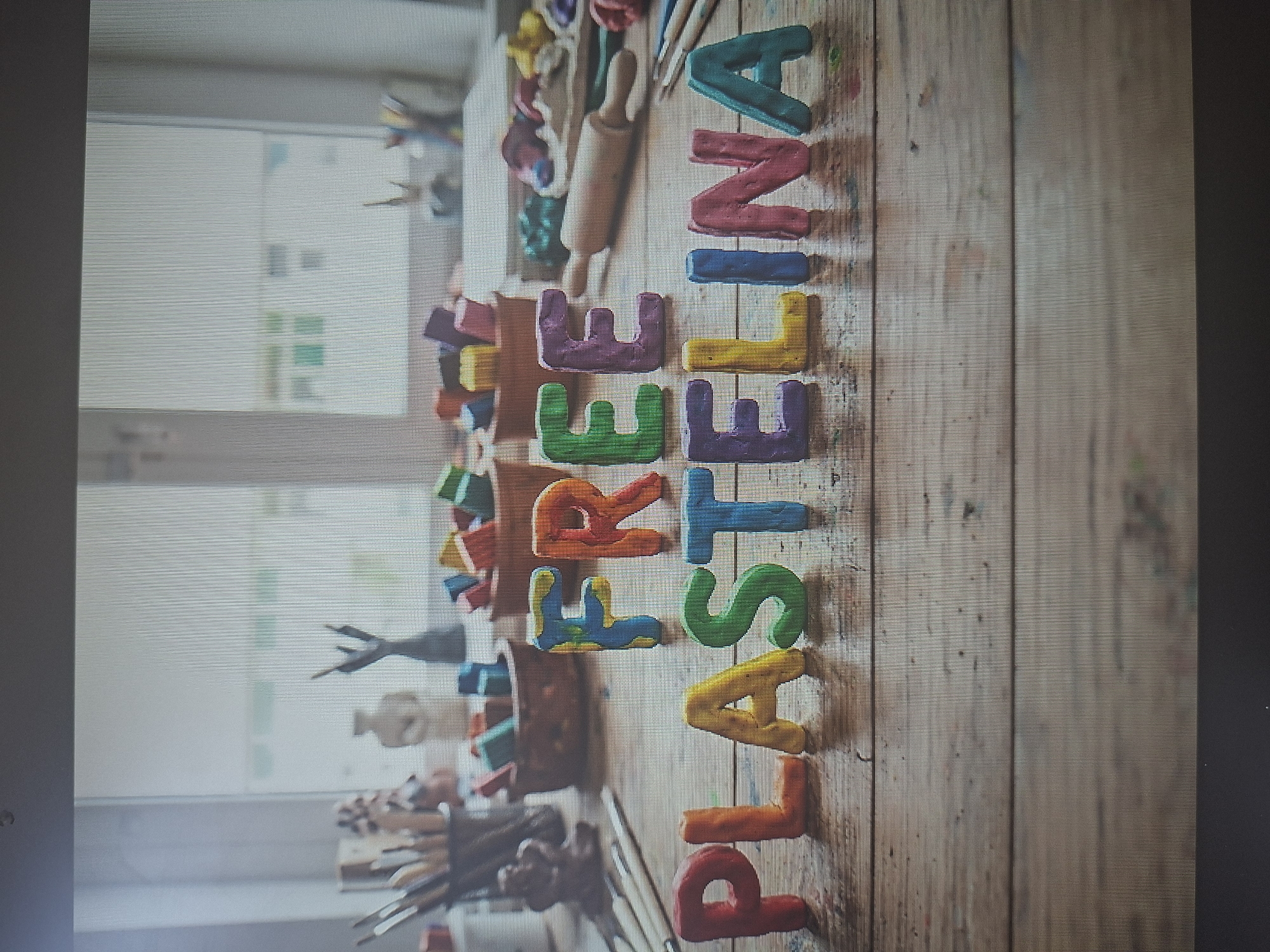 The image shows a wooden table with art supplies and the word "PLASTILINA" spelled out in colorful clay letters. In the background, a window reveals a cityscape. While the image gives the impression of being taken in an art studio with a view of Tel-Aviv, Israel, I can't confirm the exact location.