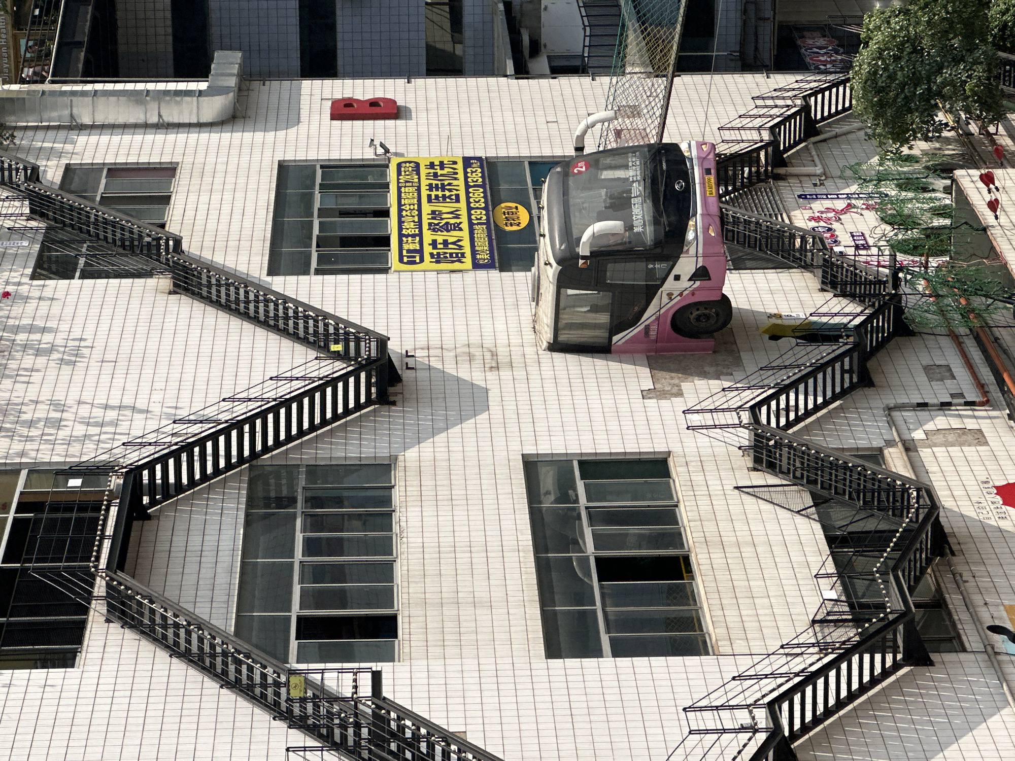 The image, taken at an unusual angle, shows the exterior of a commercial building in Nan'an District, China. The building is clad in a grid of white tiles, punctuated by rectangular windows with dark frames. Black fire escapes zig-zag up the facade on both sides. A pink and white tour bus is suspended between the two sets of fire escapes, partially obstructing a yellow advertisement sign on the building. The sign features Chinese characters and contact information. The bus displays the characters "美心文创乐园" (Meixin Creative Park) and "AMX999" and a number "2". Below the tour bus are various business advertisements on the building.
