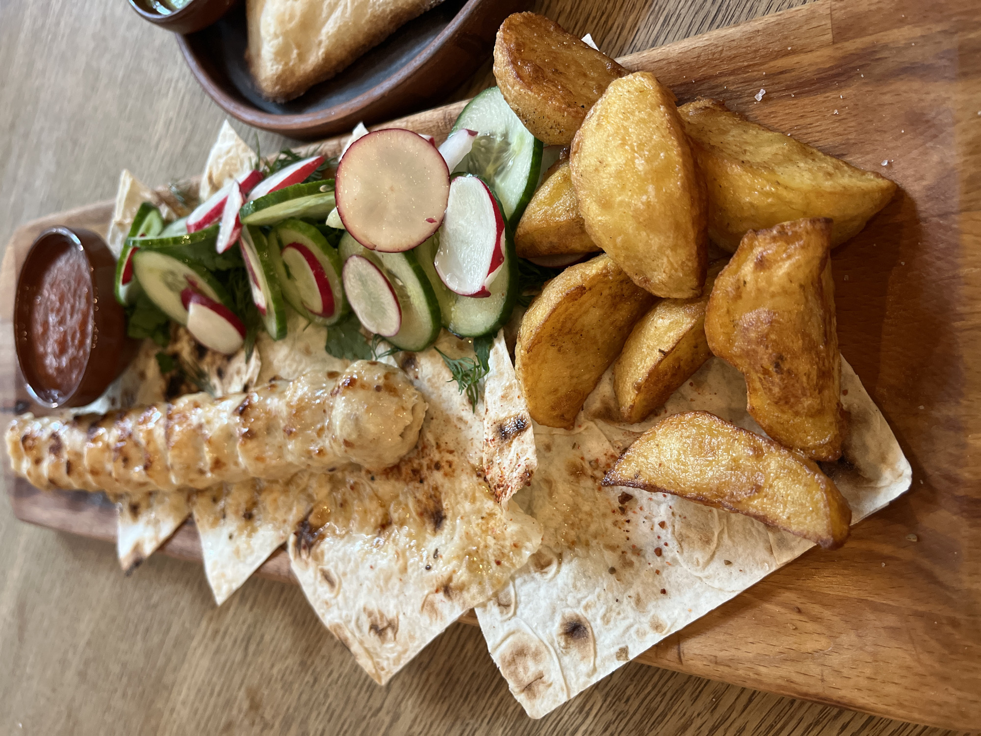A top-down view captures a platter of food artfully arranged on a rustic wooden serving board, set against a wooden table in what appears to be an indoor restaurant or cafe setting in Sochi, Russia.
The main subject is a long skewer of grilled minced meat, resembling a lula kebab, placed centrally on several pieces of thin, toasted flatbread, likely lavash. The meat has distinct grill marks and a lightly browned exterior. Alongside it are golden-brown, crispy potato wedges, scattered to the right, and a vibrant salad of thinly sliced cucumbers and round radishes, topped with fresh herbs, positioned to the left.
Two small, dark brown bowls containing sauces accompany the meal. One, on the far left, holds a reddish, possibly tomato-based sauce. The other, partially visible at the top left, contains a greenish sauce, perhaps a herb dip or tzatziki. In the blurry background, another wooden bowl with what looks like baked pastry or bread is visible.
The overall presentation is appealing, suggesting a freshly prepared meal. No people are visible, and there is no discernible text. The lighting indicates a daytime setting. The cuisine, featuring grilled meat, flatbread, and fresh vegetables, aligns well with dishes commonly found in the Caucasian region, consistent with the location context of Sochi, Russia.