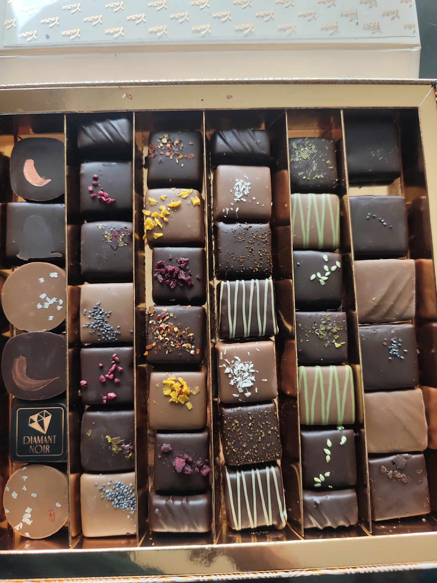 The image shows an opened box of chocolates. The box has a gold-colored interior and the lid is visible in the top part of the frame with repeated "Voisin" text printed on it. Inside the box are seven columns of assorted chocolates, each with different toppings, shapes and designs. The chocolates are separated by gold dividers. In the first column on the left, one of the chocolates bears the logo "DIAMANT NOIR."