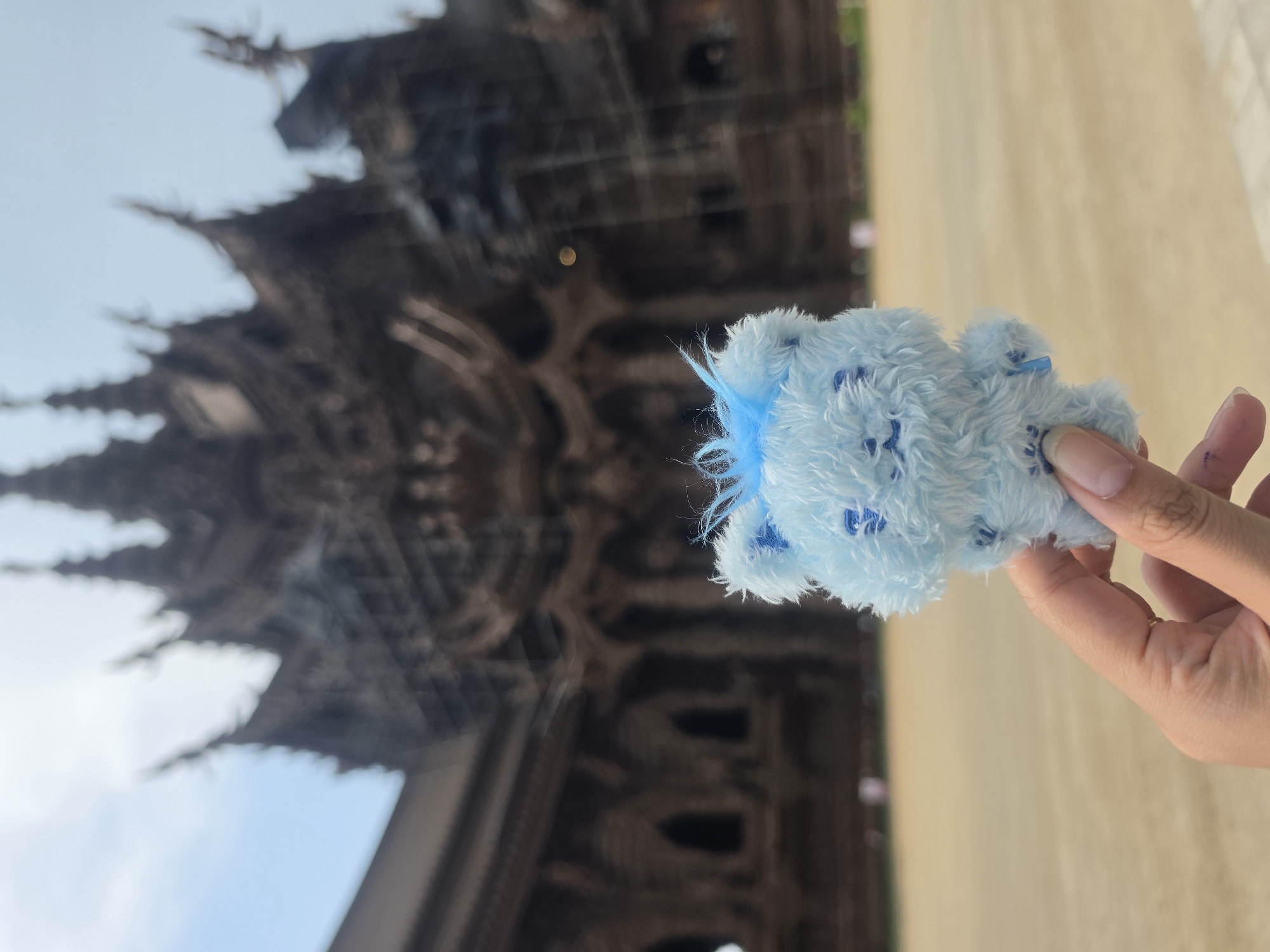 A hand holds a light blue plush bear in front of the Sanctuary of Truth, a wooden temple, in Pattaya City, Thailand. The temple is in the background, with intricate carvings visible. The sky is blue. The ground appears to be tan. The hand has painted fingernails and a ring on the finger.