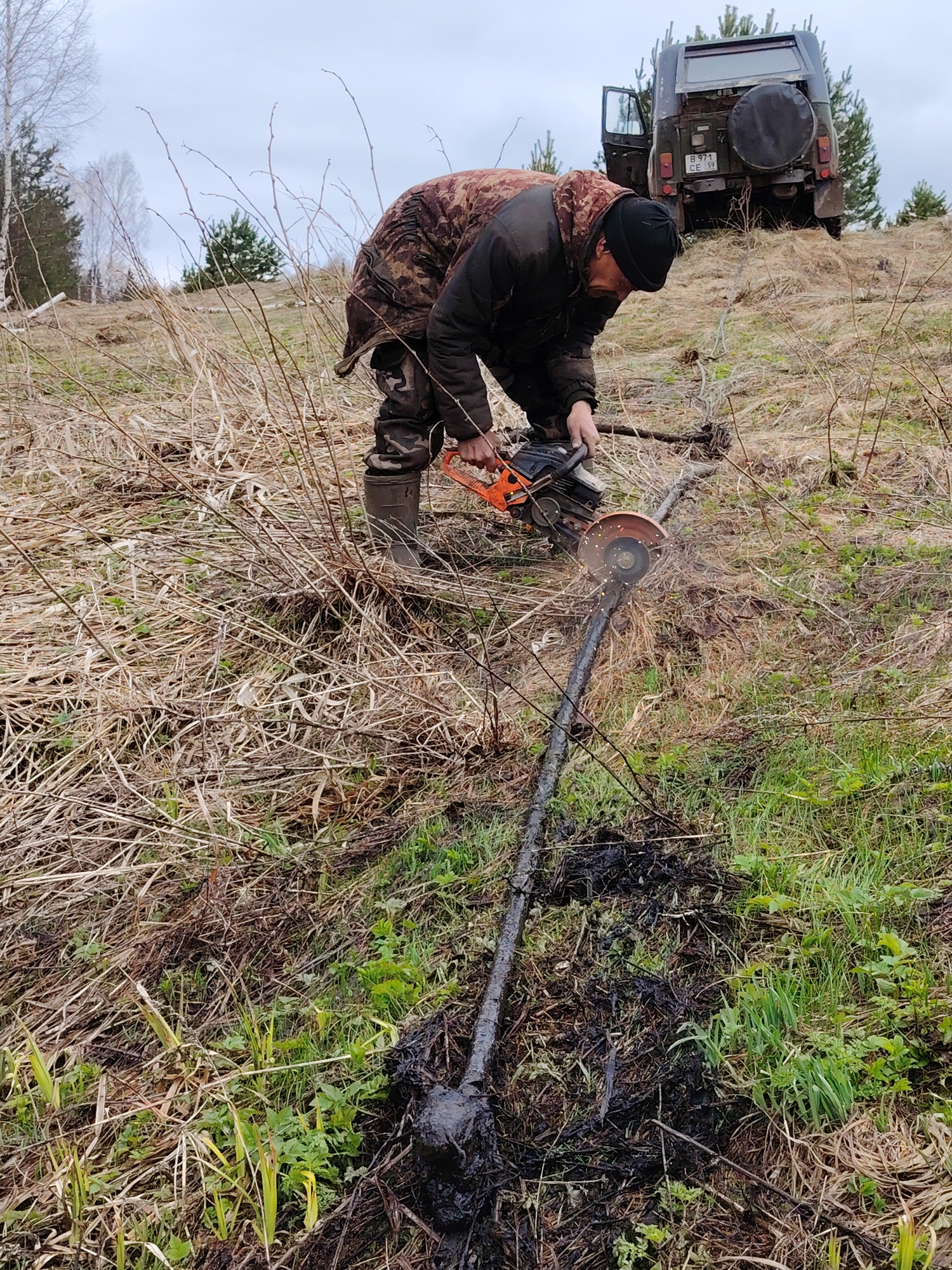 A man in Markovskii, Russia, is using a power tool to cut a long, dark metal object in a field of dry grass and green vegetation. The man wears a dark jacket, camouflage pants, dark boots, and a black cap. The metal object is partly buried in the ground, which is covered in black mud or oil. A dark green utility vehicle with an open door is parked on a small rise in the background. The sky is overcast. The license plate on the vehicle reads "B 971 CE 59".