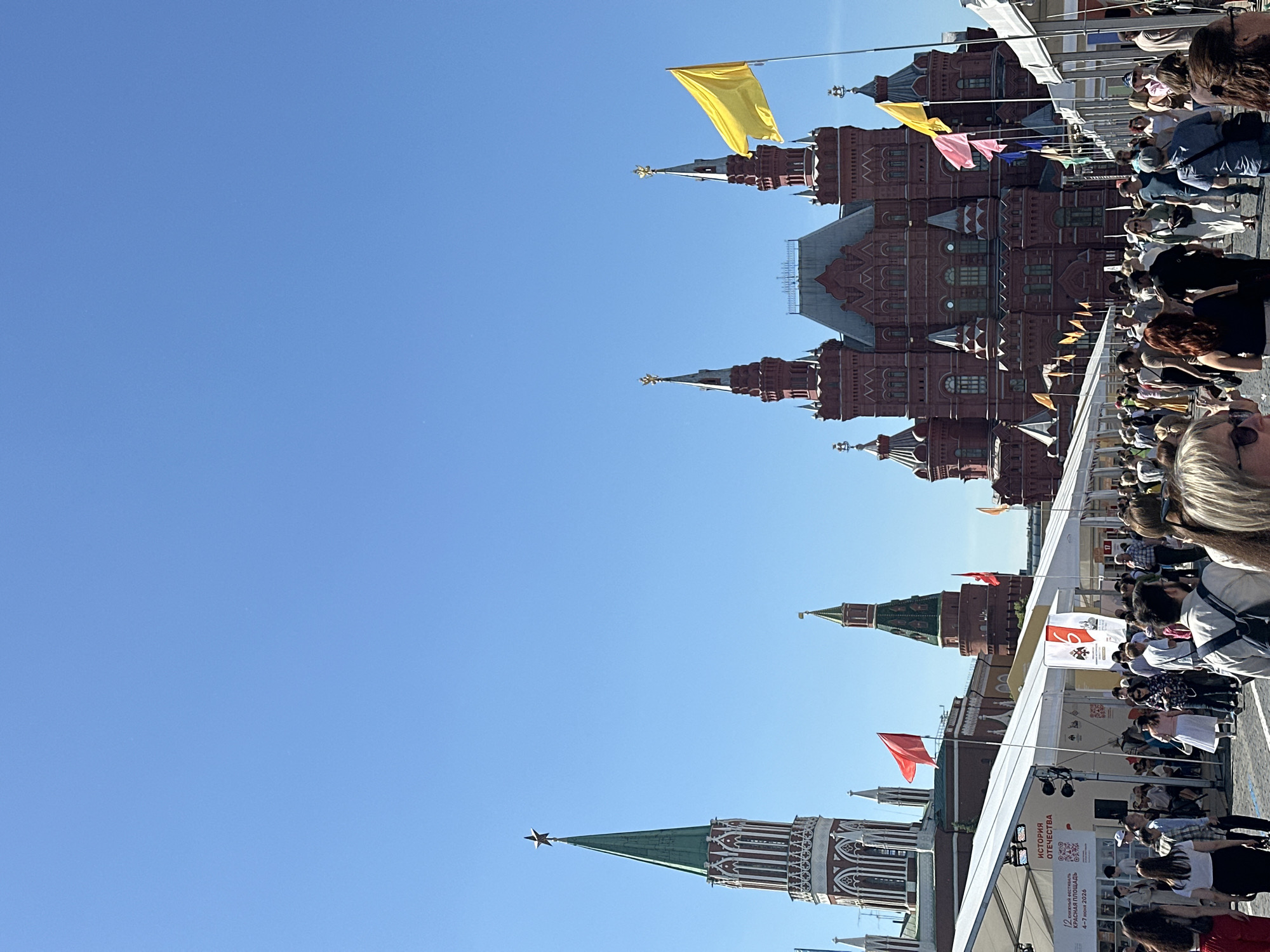 The image captures a crowded outdoor scene in Red Square, Moscow, Russia, under a clear, bright blue sky. The prominent red brick State Historical Museum, characterized by its multiple ornate spires and gold-topped finials, dominates the right side of the background. A large yellow flag is unfurled on a pole in front of the museum, with smaller pink, blue, and yellow flags visible further along. To the far left, a tall tower of the Moscow Kremlin, featuring a green conical roof and a distinctive red star, rises above other structures. Below and to the right of the Kremlin tower, another smaller red brick tower with a green roof is visible.
In the midground and foreground, a wide, paved area is lined with numerous white, modular temporary tents or stalls, indicating an outdoor event. One tent on the left displays text in Russian, "ИСТОРИЯ ОТЕЧЕСТВА" (History of the Fatherland), and identifies the event as the "12-Я КНИЖНАЯ ЯРМАРКА КРАСНАЯ ПЛОЩАДЬ 4-7 ИЮНЯ 2026" (12th Book Fair Red Square June 4-7, 2026). Red flags are mounted on poles along the tents on the left.
A large crowd of people fills the spaces between the tents and in front of the historical buildings. Individuals of various ages are present, dressed in casual summer clothing, and are seen walking in different directions, observing the stalls, and engaging with the event. Several individuals in the foreground are partially visible, including a woman with dark hair in a black top on the left and a woman with glasses and blonde/grey hair on the far left. The scene suggests a busy public gathering on a sunny day.
