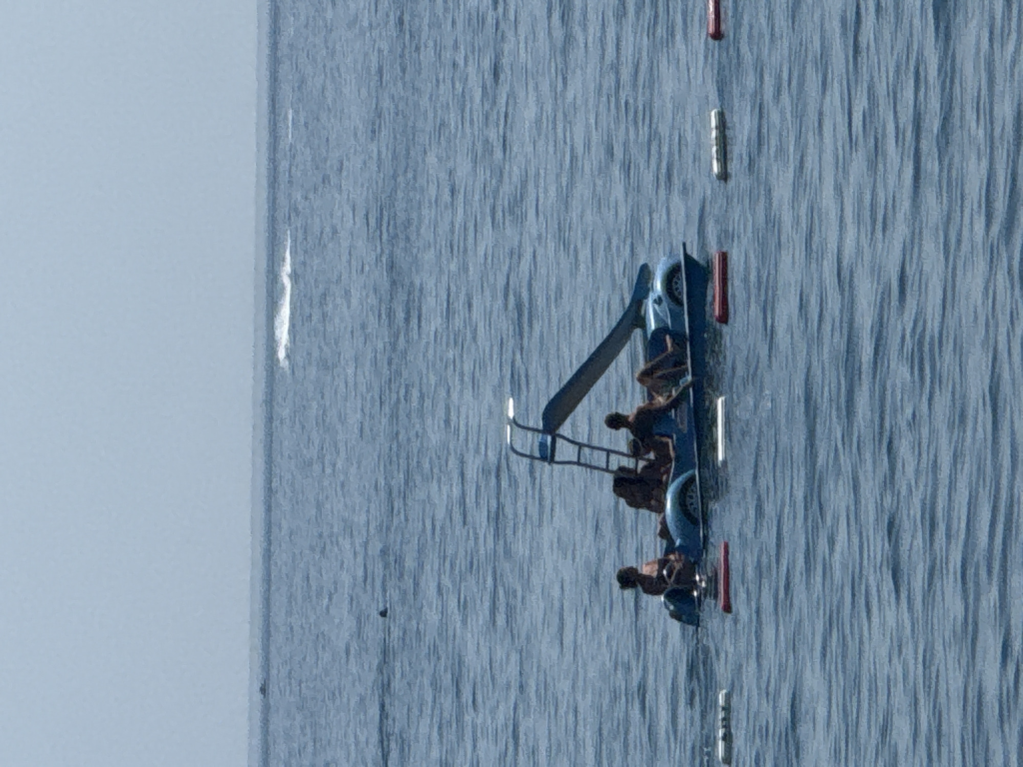 An aerial view shows a blue pedal boat with a slide on a calm sea. Four people are seated in the boat. The sky is clear and pale blue, with a hazy horizon line. Three buoys, two white and one red, are visible in the foreground of the water. The scene is calm and peaceful. It is not possible to determine the location in the city of Boreti, Montenegro from the image.