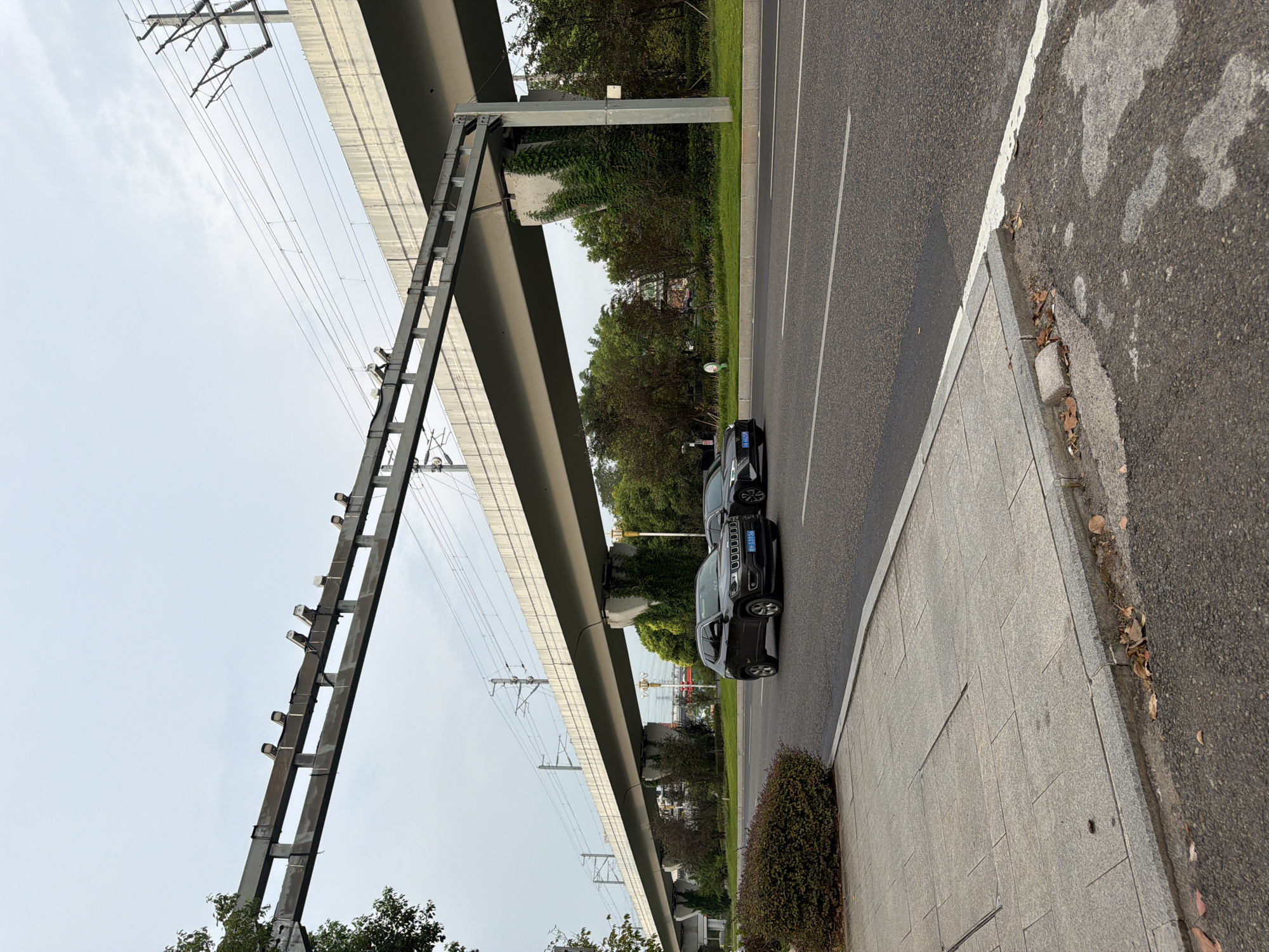 The image depicts an outdoor scene featuring an elevated roadway or railway structure. Beneath this structure, a two-lane asphalt road is visible, with two dark-colored passenger vehicles in motion. The vehicle in the foreground is a dark gray Jeep SUV, and behind it, a darker sedan is partially visible. The road is marked with white lane lines. To the left of the road, a paved sidewalk or pedestrian walkway is present, constructed of large, light-colored rectangular pavers. A neatly trimmed green hedge abuts the sidewalk. Beyond the road and to the right, there is a grassy median with trees and vegetation. The elevated structure is supported by concrete pillars, some of which are covered in green ivy. Overhead, electrical wires and pylons are apparent, suggesting a transportation corridor. The sky is overcast, indicating a cloudy day. The scene appears to be a section of urban infrastructure, possibly within the city of Jiangjin, China, given the context. The image is captured from a slightly low angle, looking up towards the elevated structure.