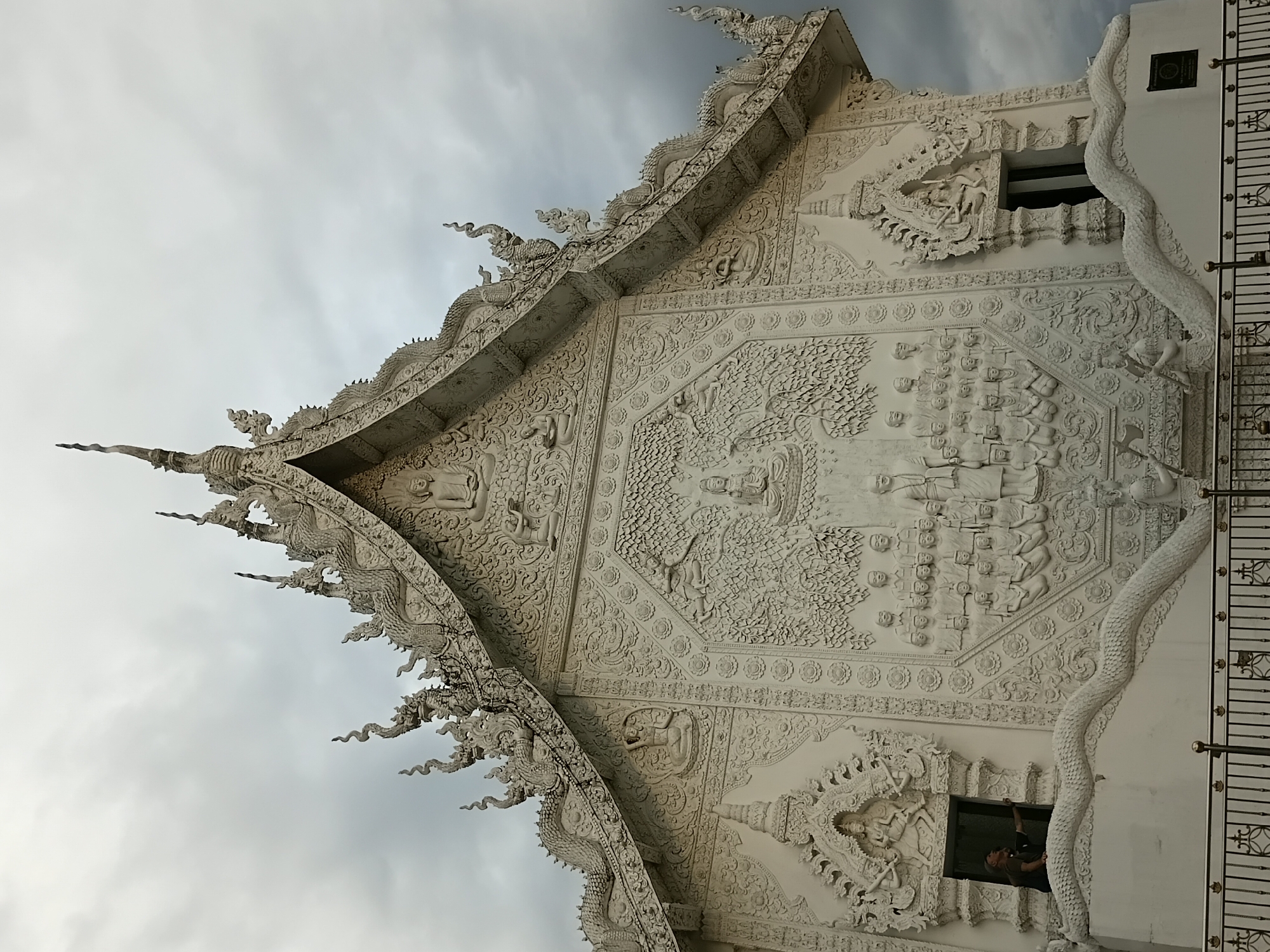 A low-angle, full shot captures the ornate white facade of a Buddhist temple in Baan Lao Phatthana, Thailand, under a cloudy sky. The temple is adorned with intricate bas-relief sculptures depicting Buddhist figures, mythical creatures, and scenes from religious narratives. Architectural elements such as tiered roofs, decorative finials, and dragon-like serpent sculptures along the entrance and roofline are prominently featured. A person is visible in the upper left window, looking outward and appearing to interact with something outside the frame. The foreground shows a black metal fence with ornate railings. The overall impression is one of grandeur and detailed craftsmanship against a dramatic, overcast sky.