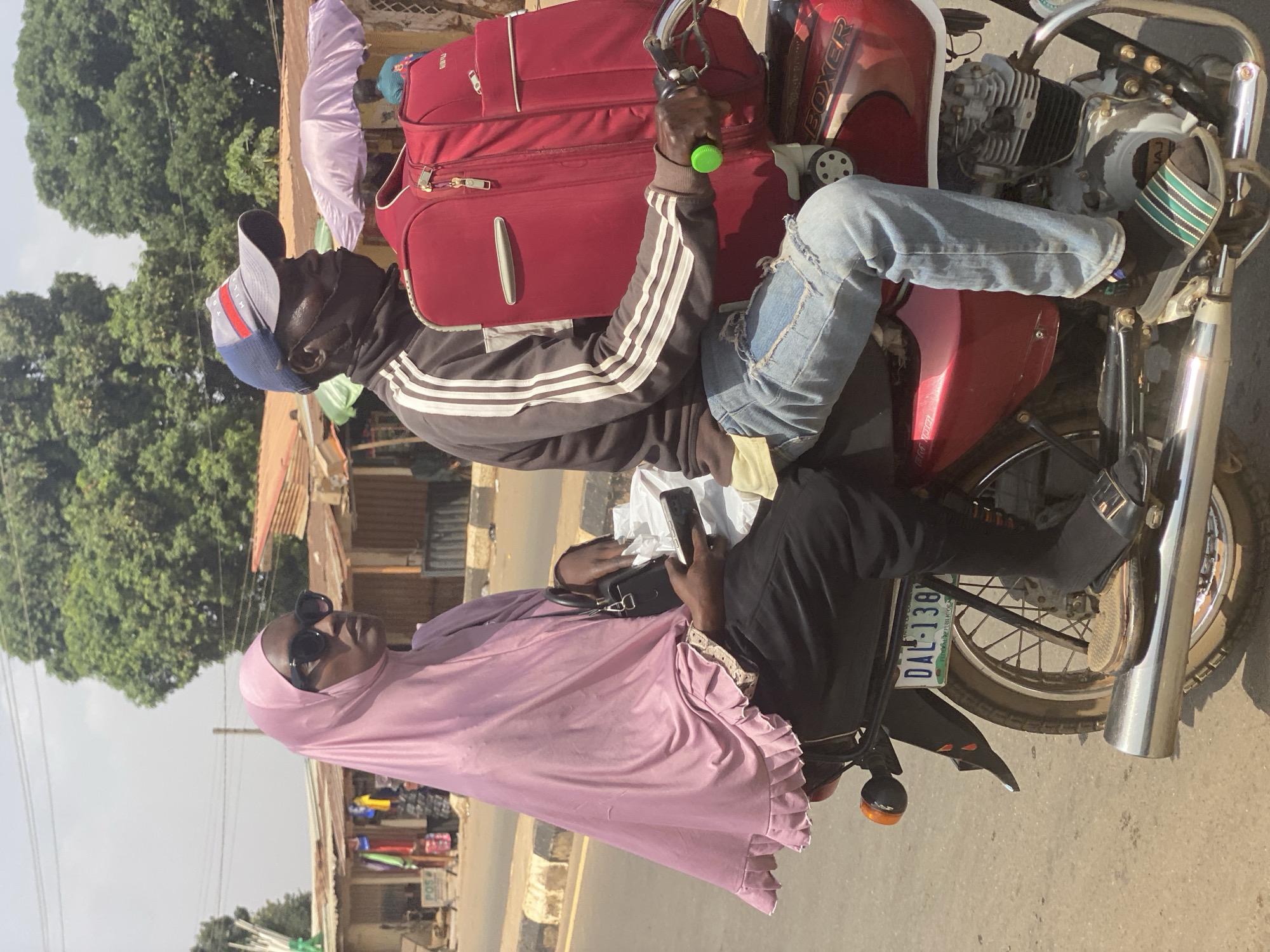 A man and a woman are riding on a motorcycle in Kafanchan B, Nigeria. The man is driving, wearing a baseball cap, a black jacket with white stripes, and blue jeans. He has a red suitcase strapped to the front of the motorcycle. The woman is sitting behind him, wearing a pink headscarf and sunglasses, and holding a cell phone and a small purse. The motorcycle has a license plate that reads "DAL-138". The background includes buildings and trees, typical of a street scene in Nigeria.