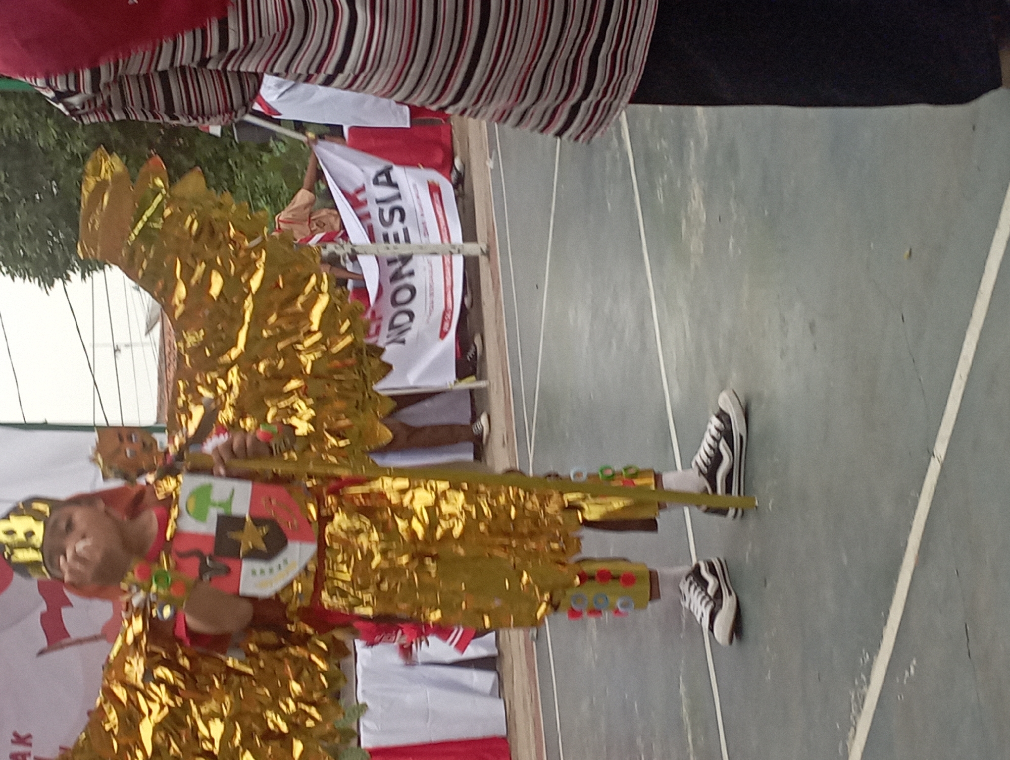 A young child, dressed in a golden costume resembling a bird with large wings, stands on a grey court surface. The child is wearing a gold crown with the number "19" on it and a red shirt with a coat of arms on the chest. The costume includes golden, crinkled material for the wings and pants, adorned with colorful circular decorations on the legs. The child is holding a golden staff and appears to be in motion, possibly performing. In the background, a banner with "NDONESIA" printed on it is visible, along with several people in casual attire and what appears to be a school or community event setting. The overall scene suggests a festive or cultural celebration.