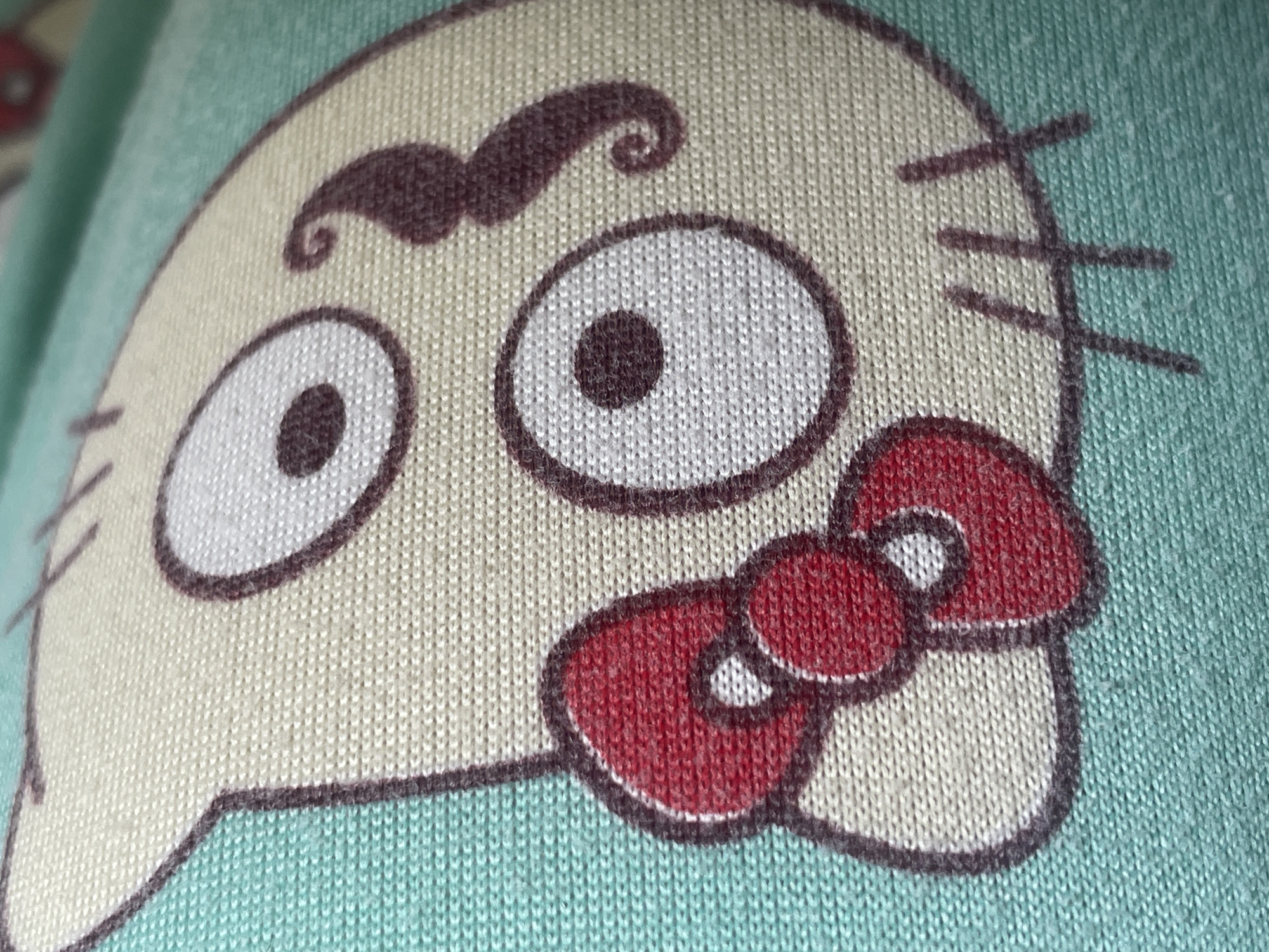 The image is a close-up, high-angle shot of a fabric with a printed cartoon image of a skull-like character. The character has a cream-colored face with large white eyes with dark pupils and thick black outlines. It has a red bow tie on the left side of its face and a curly, dark brown mustache above its nose area. The background is a textured turquoise fabric. The image does not contain any elements that would allow for its location to be identified in Tashkent, Uzbekistan, or any other city.