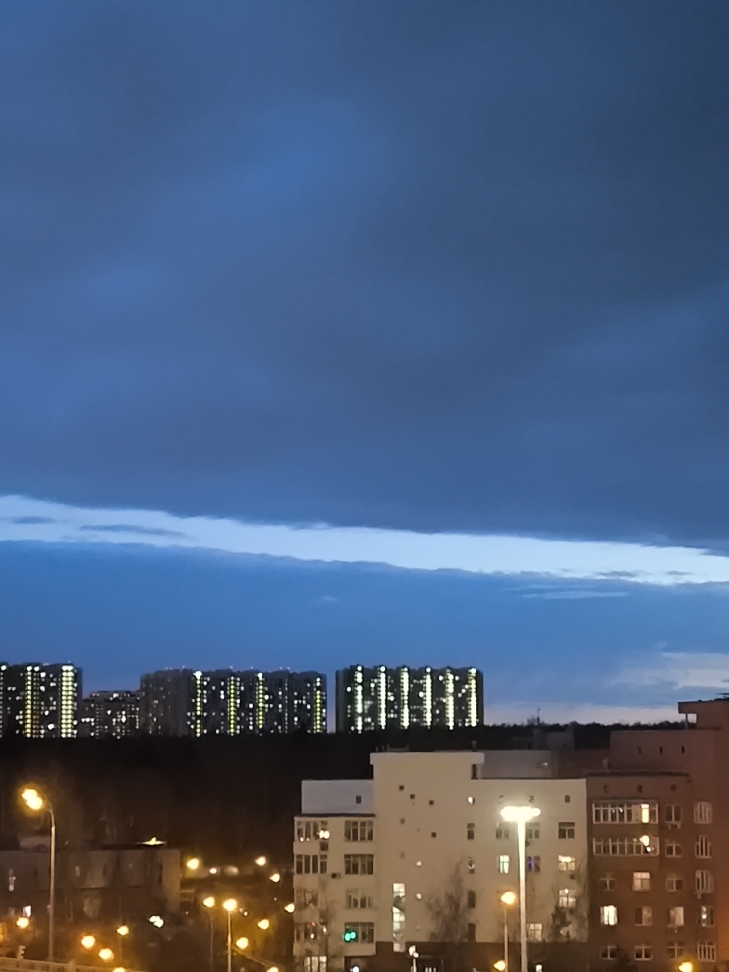 The image depicts a cityscape at night, likely in Khimki, Russia. The sky is a dark blue, with a thick band of clouds towards the horizon. The skyline features several high-rise apartment buildings, illuminated with rows of lights, indicating occupied units. In the foreground, there are shorter residential buildings, also lit. Streetlights cast a warm, yellow glow on the scene. The photo suggests a quiet urban environment in the evening. There are no people visible in the frame.