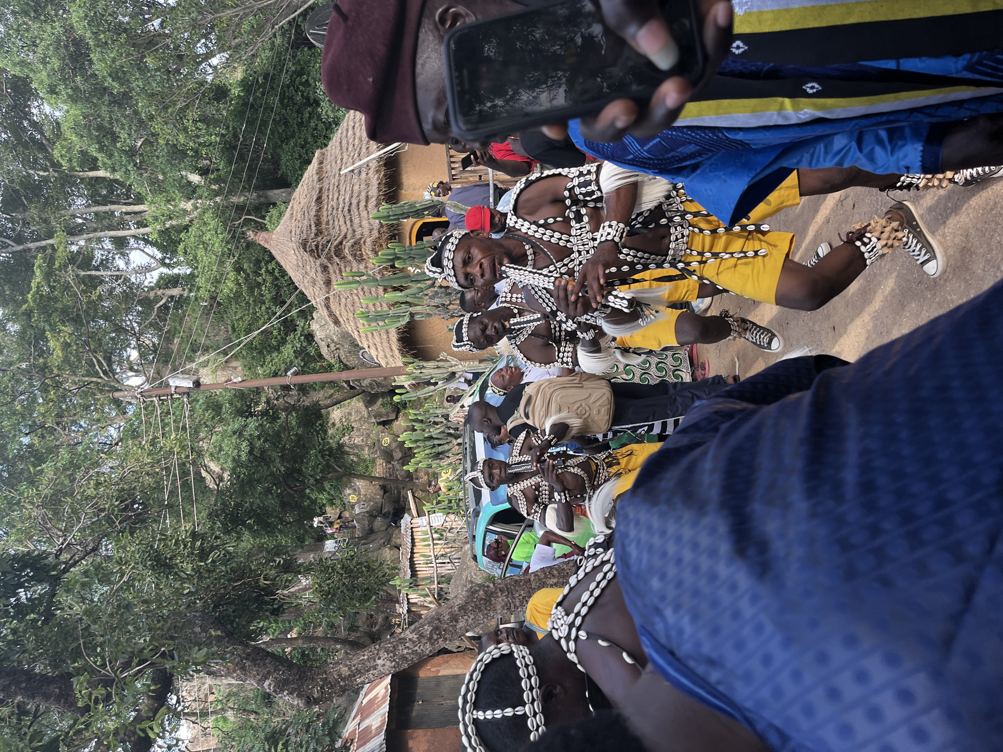 The image shows a group of men in traditional attire, some playing musical instruments, at an outdoor event in Jos, Nigeria. The man in the center is playing a flute-like instrument and is adorned with shell necklaces and headdresses. He is wearing yellow shorts and dark sneakers. Others in the foreground also wear similar shell adornments. The background features lush greenery, a thatched-roof structure, and a vehicle. Some onlookers are present, with one person in the immediate foreground holding a smartphone. The overall scene depicts a lively cultural gathering.