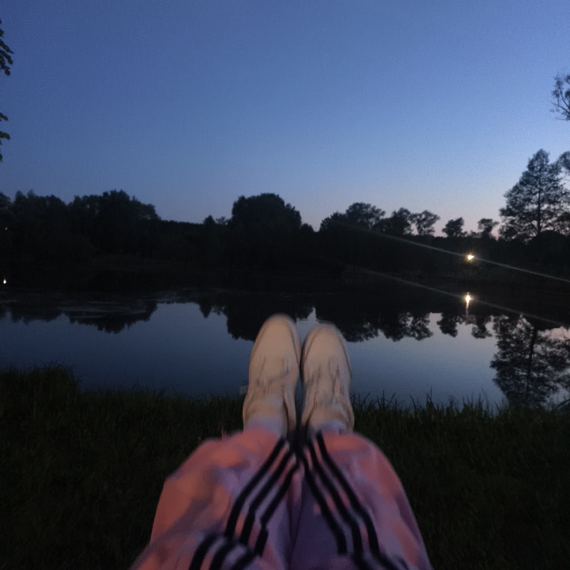 A low-angle, eye-level photograph taken at dusk in what appears to be a natural environment, possibly a lake or river bank. In the foreground, there is a partial view of a person wearing light-colored sneakers and pink athletic pants with black stripes. The person appears to be seated with their legs extended towards the body of water. Behind the person is a body of water with the reflection of the treeline visible in the water. The treeline appears to be dense with trees. Above the treeline is a gradient sky with lighter shades of blue near the horizon and darker hues toward the top of the image. A pole, likely a fishing rod, is barely visible extending over the water's surface and a light source is present to the right, in the trees. This is not necessarily in Варзи-Ятчи, Russia. The exact location within that region is not possible to determine from the available visual information.