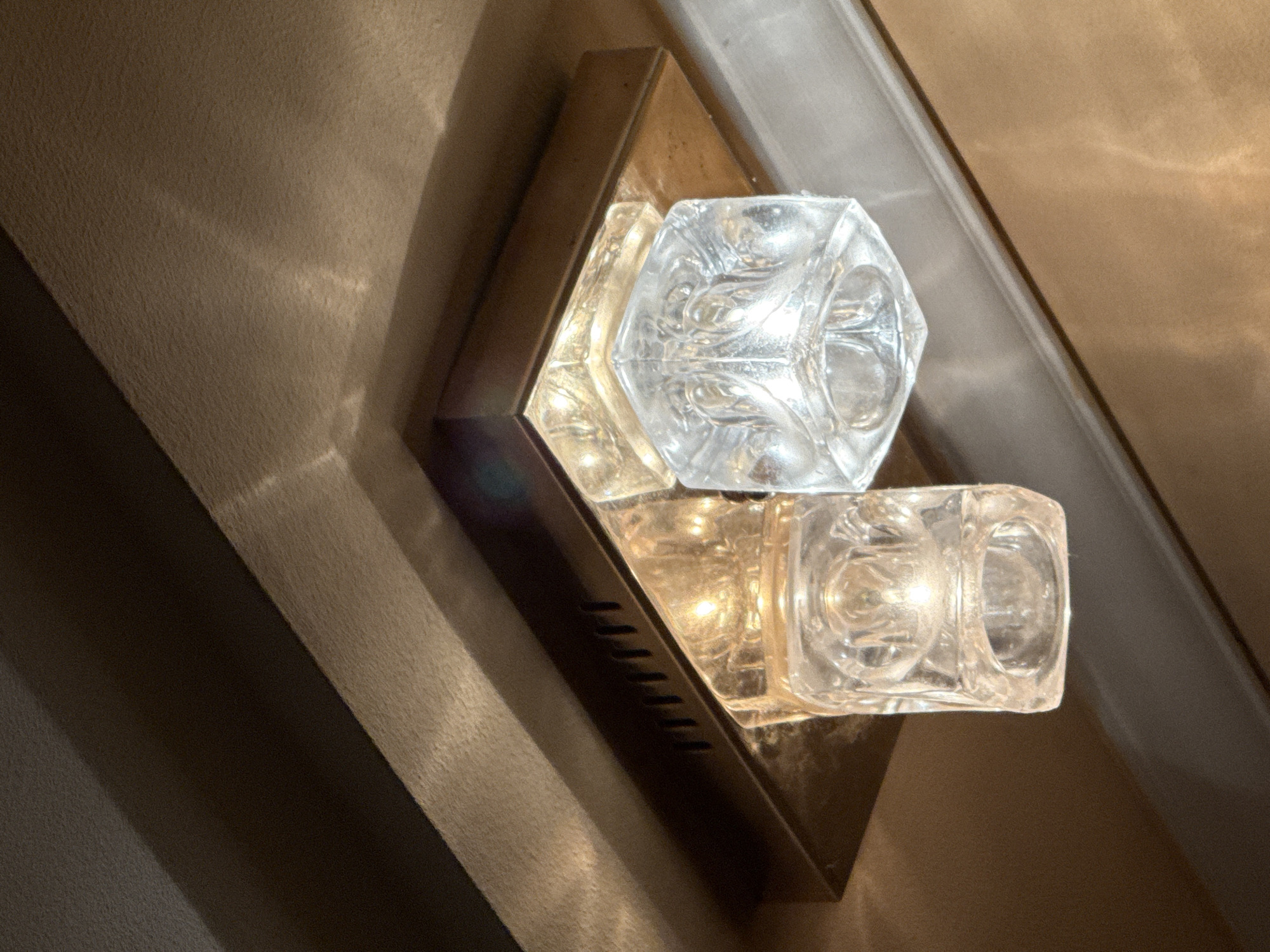 A close-up, low-angle shot focuses on a modern ceiling light fixture featuring two square glass shades. The shades have a textured, etched pattern and are mounted on a dark bronze or brass rectangular base. One shade emits a warm, yellowish light, while the other emits a brighter, whiter light. The fixture is attached to a textured ceiling with recessed panels. The background consists of angled surfaces, suggesting an interior architectural detail. There are no people or animals present in the image. This image was captured in the city of Okhtyrka, Ukraine.