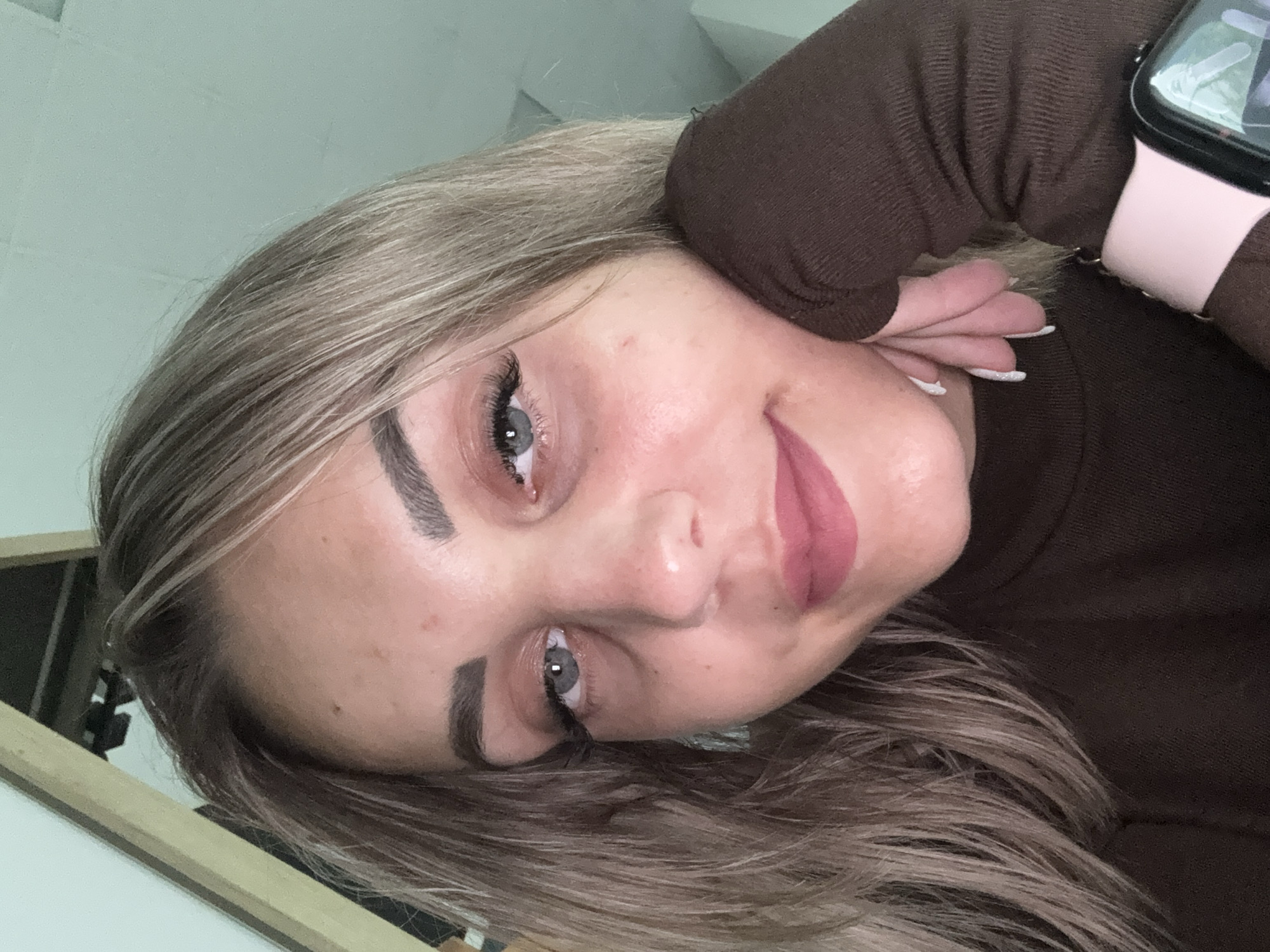 A young woman with blonde hair is pictured in a close-up selfie. She is wearing a brown turtleneck and a pink smartwatch. Her left hand is resting on her cheek. The background appears to be an indoor setting with a light green wall and some wooden trim. There are no visible indications of this scene being in Yuzhno-Sakhalinsk, Russia.