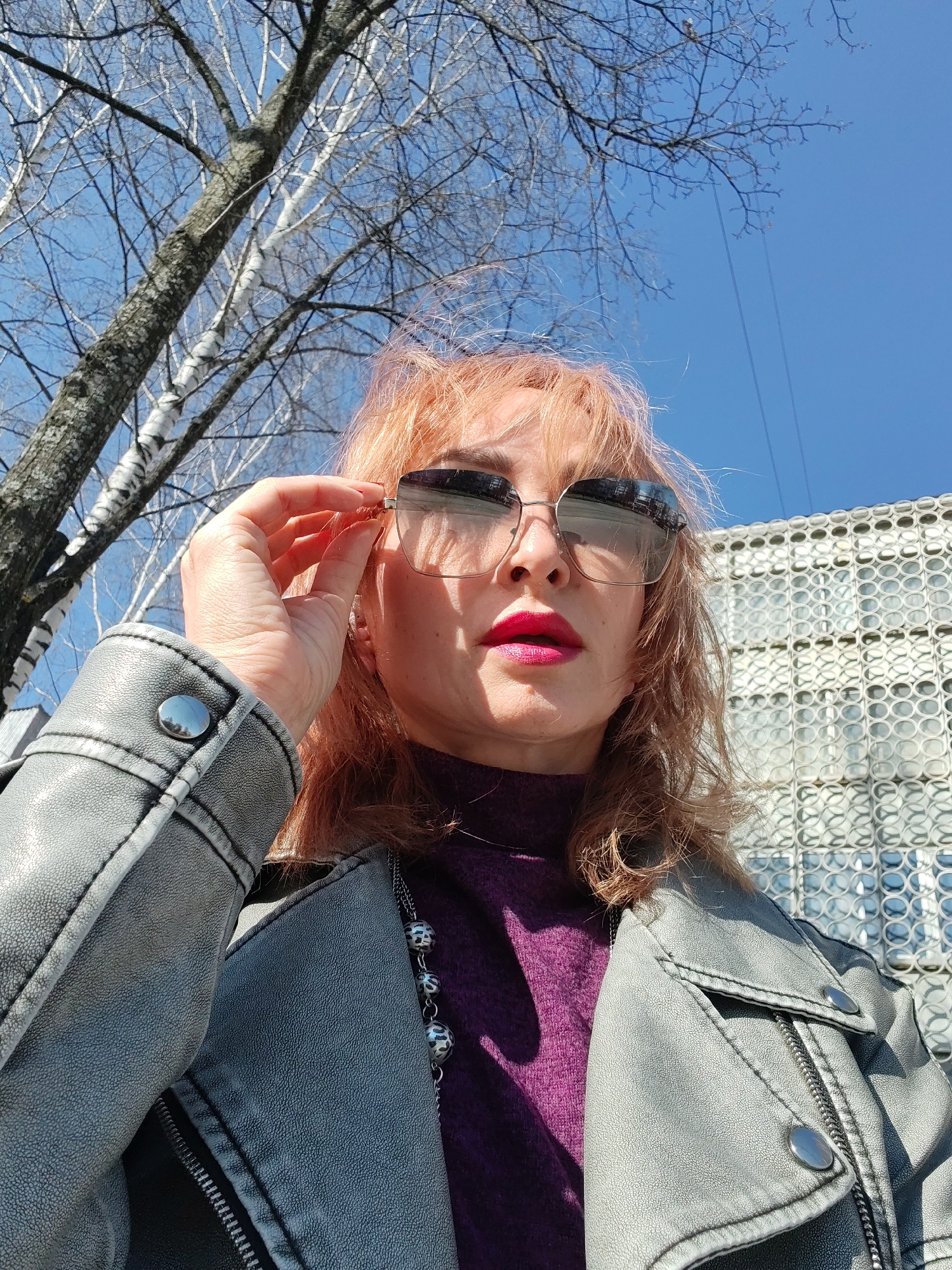A medium, low-angle shot depicts a fair-skinned woman with dyed reddish-blonde hair and bright fuchsia lipstick standing outside. She is wearing sunglasses and a purple turtleneck sweater, partially covered by a grey leather jacket with silver buttons and zipper. A silver necklace with patterned beads is visible. Her right hand is raised, adjusting the sunglasses.
Behind her to the left is a bare birch tree with a blue sky visible between its branches. To her right is a building with a facade of circular decorative elements. The location is Chaikovsky, Russia.