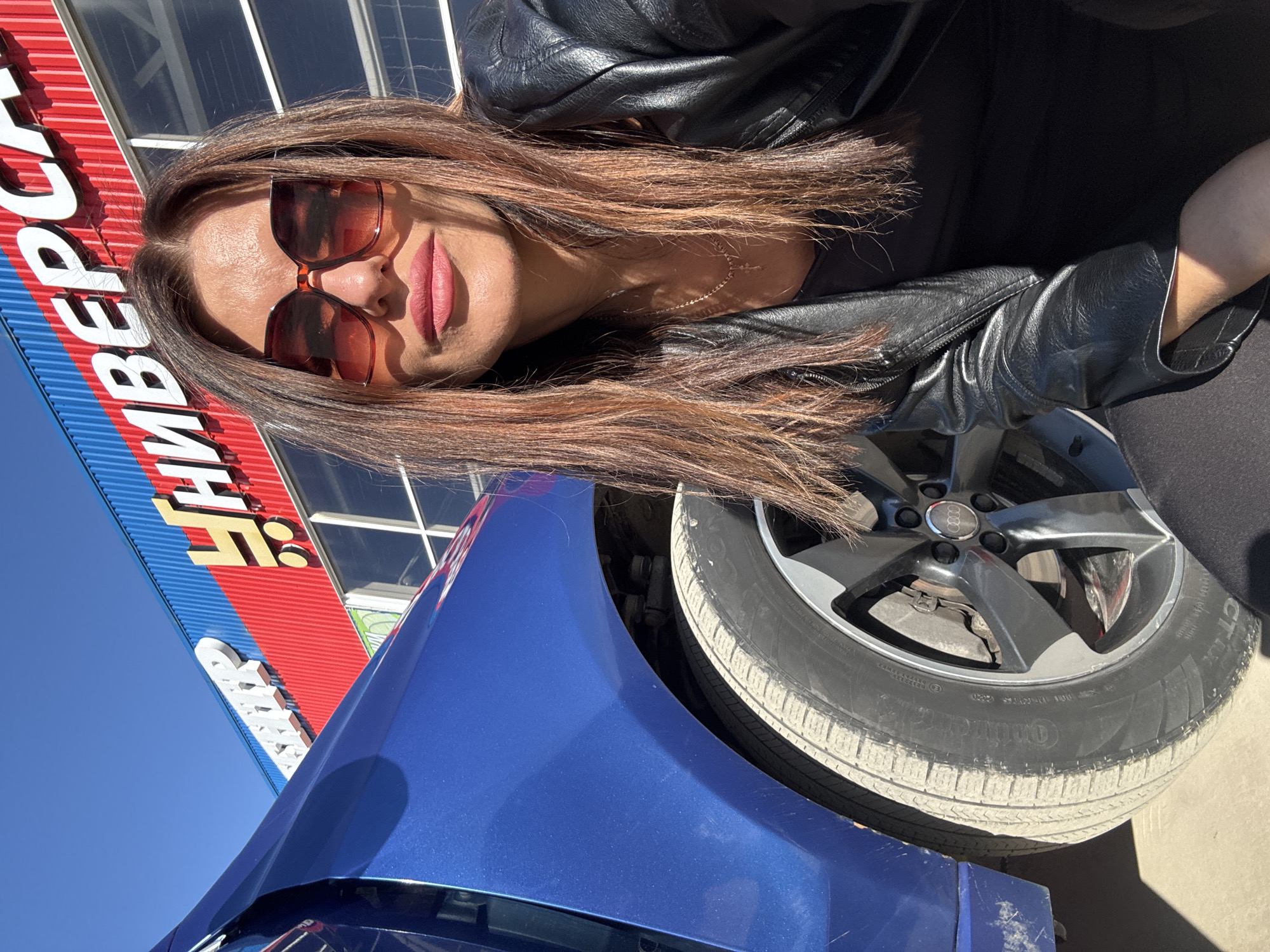 The image is a medium shot of a woman wearing sunglasses and a black leather jacket, positioned next to a blue car. The car's tire is visible and appears to be a Continental brand. In the background, there is a building with a sign written in Russian, "УНИВЕРСАМ", indicating a universal store. Based on the language and the building's appearance, the location is likely in the city of Chaikovsky, Russia. The woman is facing the camera with her head tilted. The sky is clear blue.