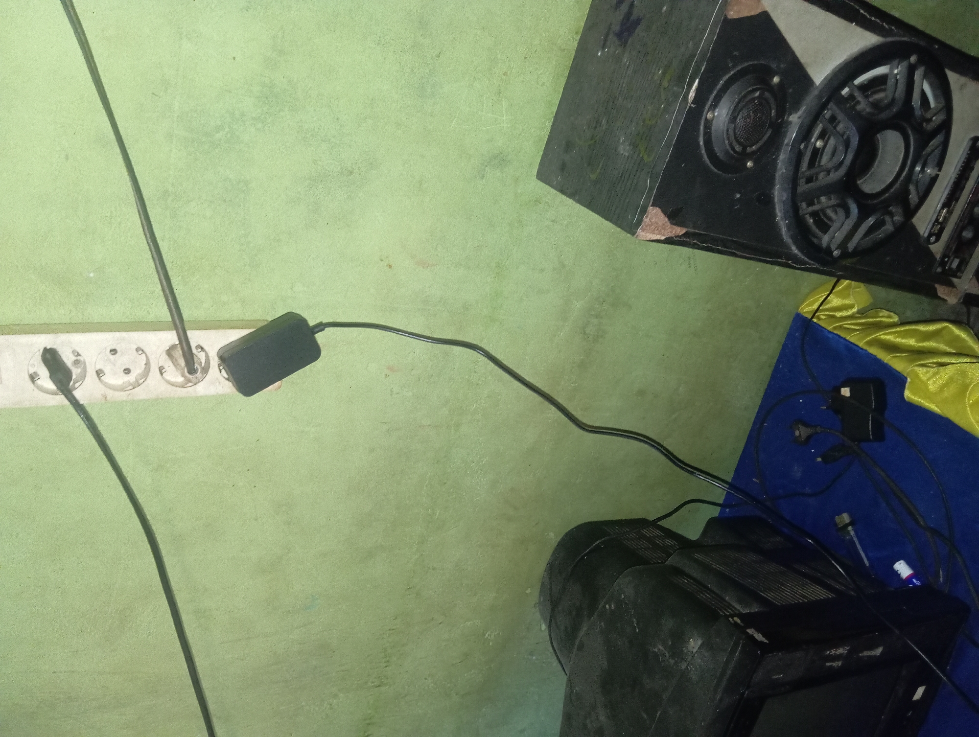 In Rejosari, Indonesia, an image depicts a corner of a room with a pale green, textured wall. A white electrical outlet strip with three circular sockets is mounted on the wall. A black power adapter is plugged into the top socket, with its black cable extending downwards and then diagonally upwards, connecting to an unseen device. To the left, a black television set sits on a blue surface. To the right, a tall, black speaker cabinet with visible damage to its edges is positioned on the floor. Various black cables and power adapters are present around the television. The overall scene suggests a functional, though somewhat cluttered, electronic setup.