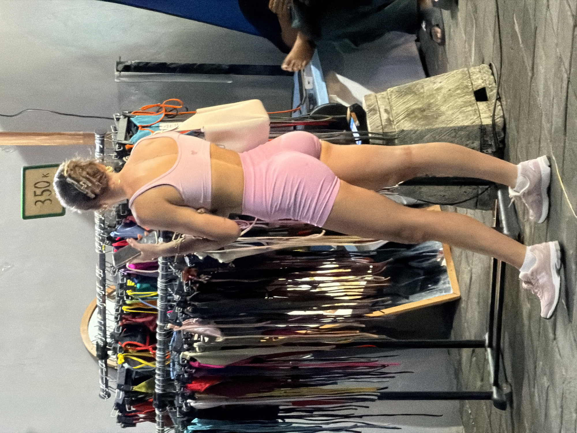 A woman in a light pink workout outfit and pink sneakers stands at a clothing rack in what is likely a retail environment. She appears to be looking at or interacting with items on the rack. The location is assumed to be in North Kuta, Indonesia based on the price tag.