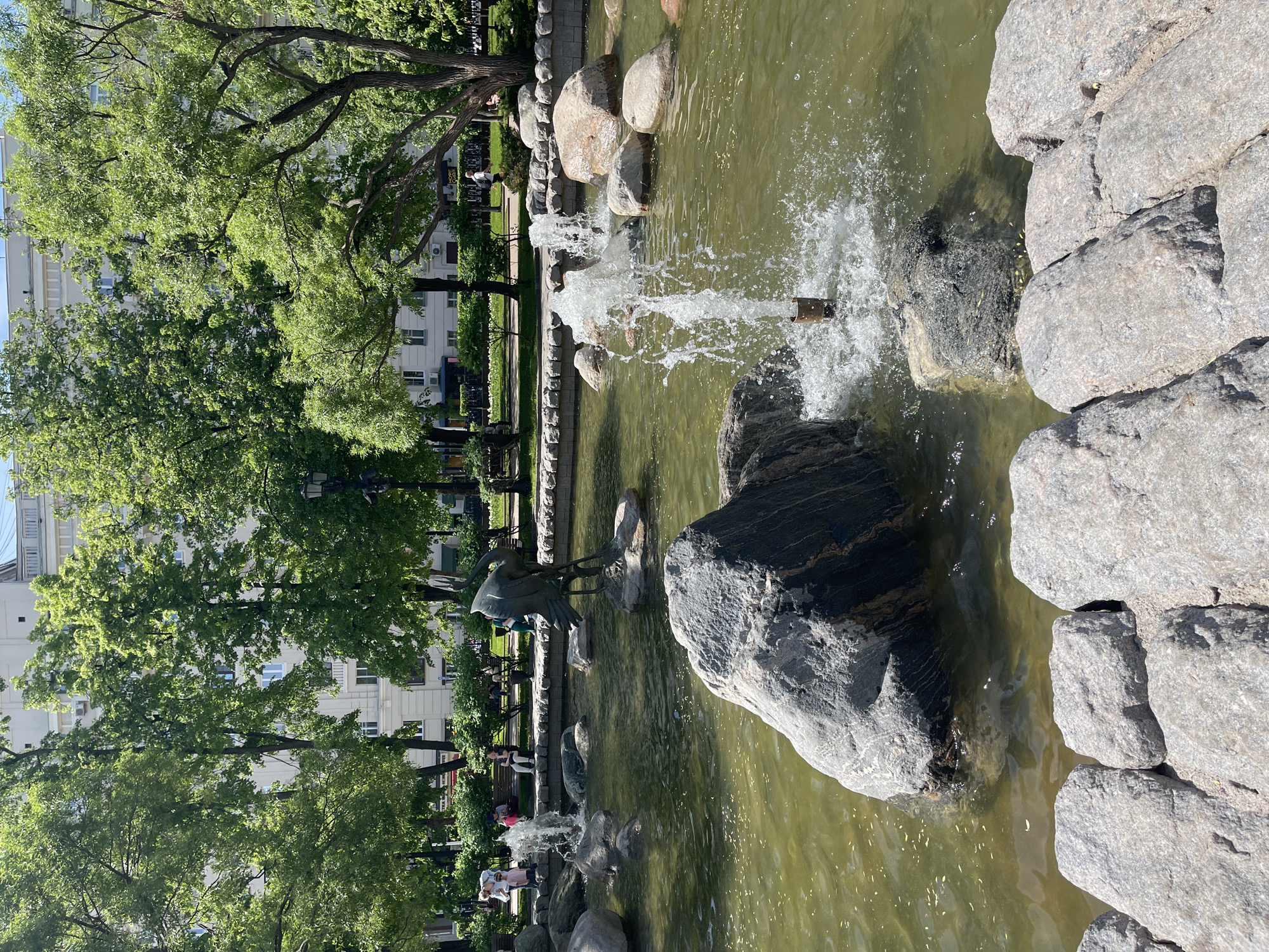 A serene urban park scene unfolds around a pond adorned with rocks and trees. The water features jets of water creating small fountains. A bronze heron statue stands near the edge of the pond. Trees and benches line the water’s edge on the left, and people are seated on the benches. Buildings are visible in the background.