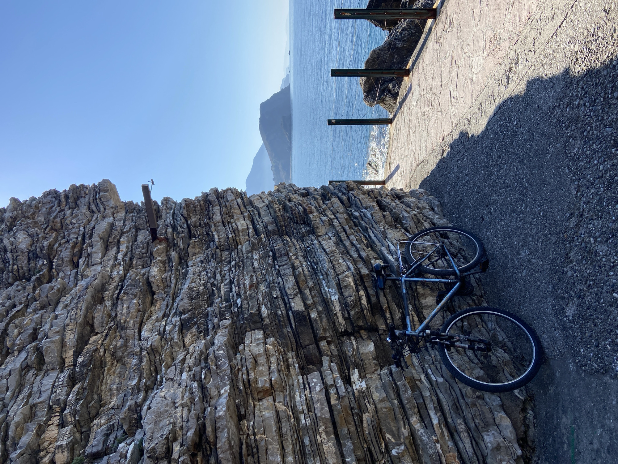 The scene is a rocky coastal area in Budva, Montenegro, featuring a light-colored bicycle propped against a large rock formation with distinct horizontal layers. The rock formation occupies a significant portion of the left side of the frame. To the right, there is a paved path or walkway, transitioning into a pebbled area. Beyond the path, the Adriatic Sea stretches into the distance under a clear blue sky. A portion of a metal barrier is visible along the edge of the path, likely serving as a safety railing. In the background, there are distant mountains or landmasses. The overall lighting is bright and sunny.
