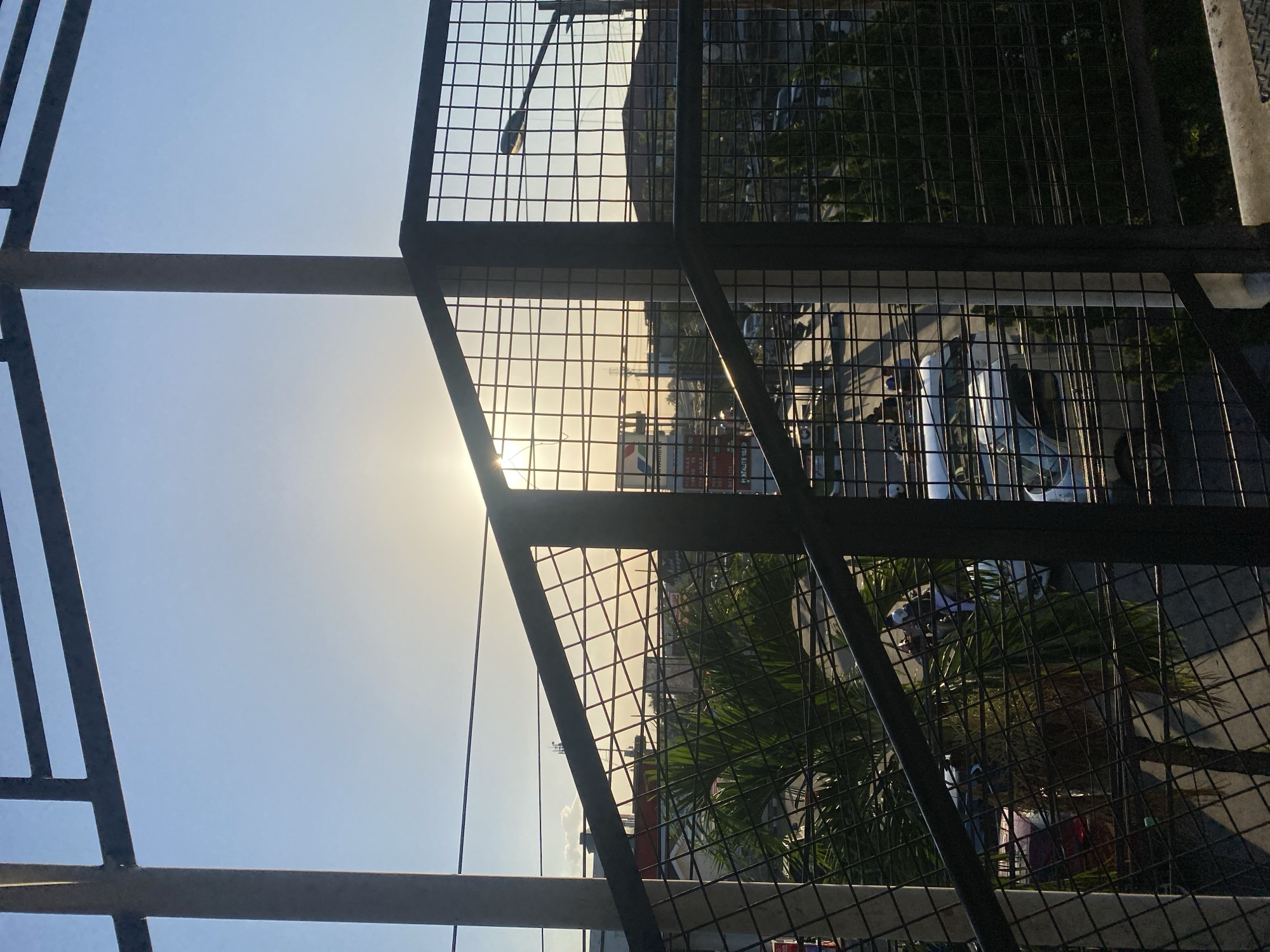 The image depicts a view through a metal grid fence, looking out onto a street scene during what appears to be late afternoon or early evening. The sun is low in the sky, casting a warm glow and creating a bright haze in the upper right quadrant of the frame. In the foreground, a concrete pillar and dark metal railing are visible, supporting the grid structure through which the scene is viewed.
Below the fence, a street is visible with several vehicles, including cars and what appears to be a van. The traffic seems moderate. A tall palm tree with lush green fronds is positioned to the left, partially obscuring the view of some of the vehicles. Several people are also visible, some walking and some appearing to be on or near motorcycles.
In the background, buildings and structures typical of a city are present. Some have signage, though they are not clearly legible. A street lamp is visible on a pole, illuminated by the setting sun. The sky is a clear, pale blue, transitioning to a warmer tone near the horizon where the sun is located.
This scene is identified as being in Banjarbaru, Indonesia, suggesting an urban environment characterized by tropical vegetation and the presence of a busy street. The overall impression is one of an everyday urban landscape captured during a picturesque sunset.