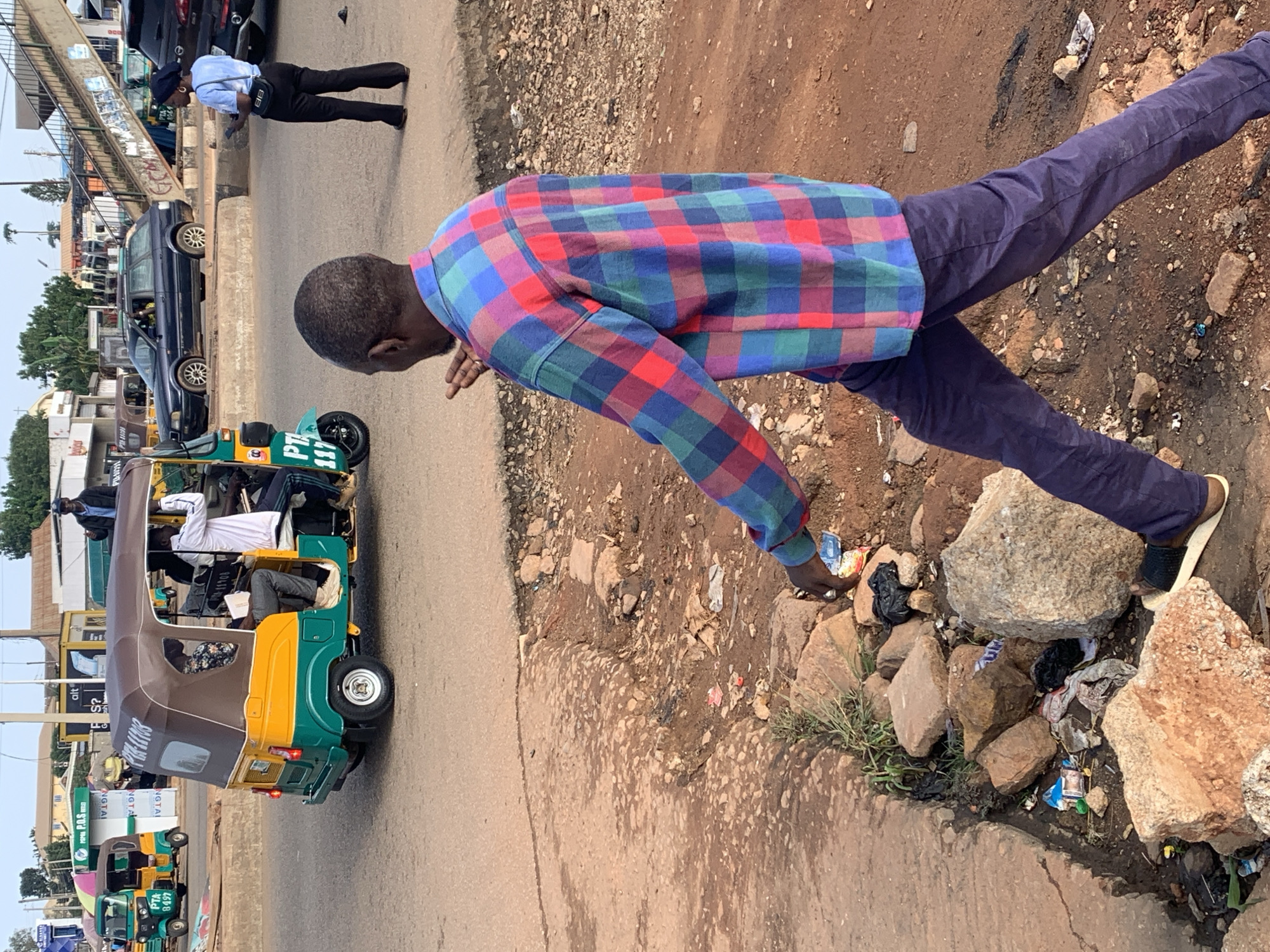 The image shows a street scene in Gura Top, Nigeria. In the foreground, a man wearing a colorful plaid shirt and dark pants is walking away from the camera, with his left hand extended outwards as if signaling. He is walking on a dirt path next to a broken concrete curb, which is littered with rocks and trash, including plastic bags and wrappers.
In the middle ground, a yellow and green auto-rickshaw, identified by the license plate "PTA 11103", is parked on the street. Several people are visible inside the auto-rickshaw. To the left of the auto-rickshaw, another similar auto-rickshaw is parked.
In the background, there are several buildings and shops, as well as a pedestrian overpass. A dark blue station wagon is parked on the street to the right of the auto-rickshaw. A woman in a blue shirt and dark pants is standing on the sidewalk, holding a mobile phone. Further in the background, other vehicles and people are visible. The environment appears to be an urban setting with a mix of paved and unpaved surfaces, and a visible presence of litter.