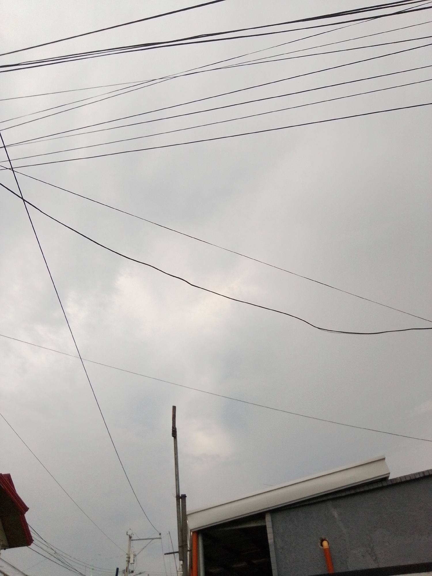 The image shows a view of the sky with numerous overhead wires crisscrossing. In the lower part of the frame, a building with a gray facade and a white overhang is visible, along with a tall wooden utility pole. To the left, a portion of a building with a red roof can be seen. The sky is overcast with clouds, suggesting a cloudy or possibly rainy day. The scene appears to be in an urban or suburban setting within Davao City, Philippines, indicated by the characteristic utility poles and building structures common in the region. There are no people or discernible actions depicted in the image.