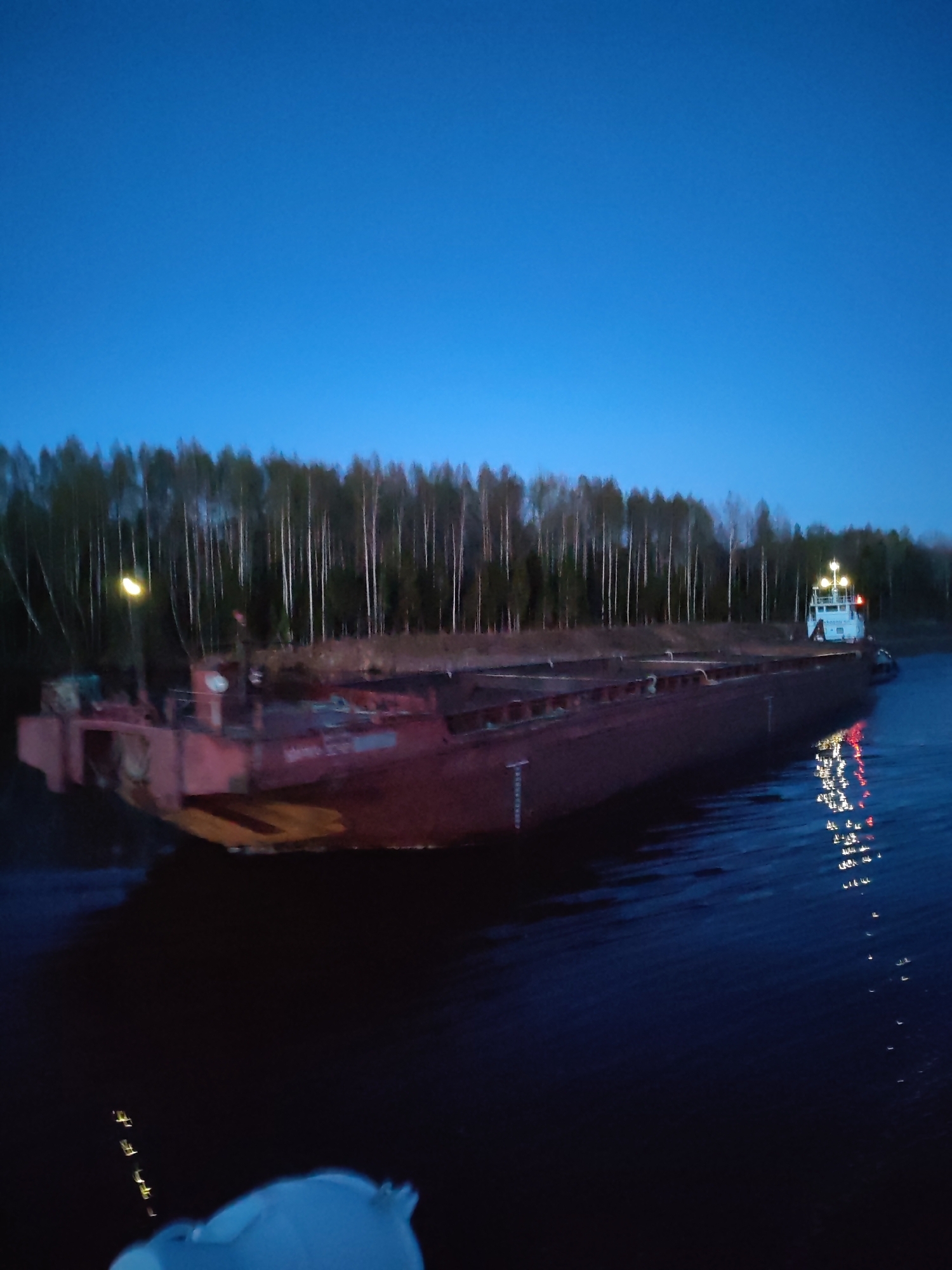 The image depicts a large, likely rusty, cargo barge on a dark body of water. The time of day is either late evening or early morning as the sky is a deep blue and visibility is low. A line of trees is visible in the background. The barge is illuminated by two lights, one on the front left and one at the rear. Reflections of the rear light can be seen in the water. The image is somewhat blurry, especially in the foreground and background. There is no specific evidence in the image to confirm the location as Uzhla, Russia.