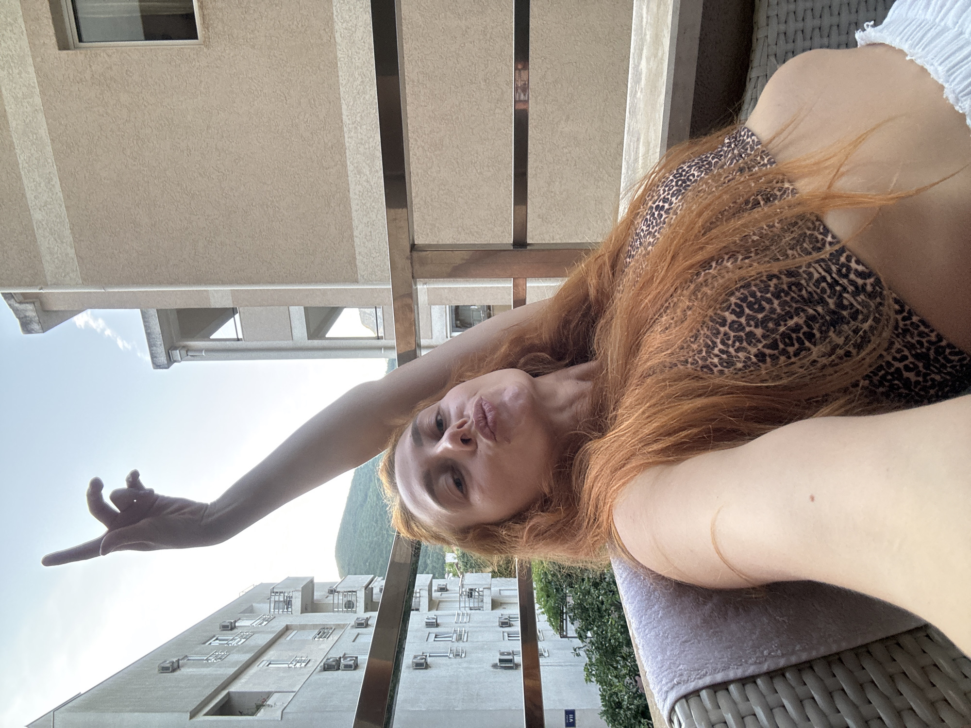 A woman with long, auburn hair, wearing a leopard print bikini top and white shorts, is photographed from the chest up on a balcony in Budva, Montenegro. She is seated on a wicker chair, leaning back and looking towards the camera with her lips pursed. Her right arm is raised overhead, with her index finger pointing upwards. In the background, a white apartment building with multiple balconies and air conditioning units is visible, along with a beige building and a distant green hillside under a clear sky. A metal railing runs across the mid-ground of the image.