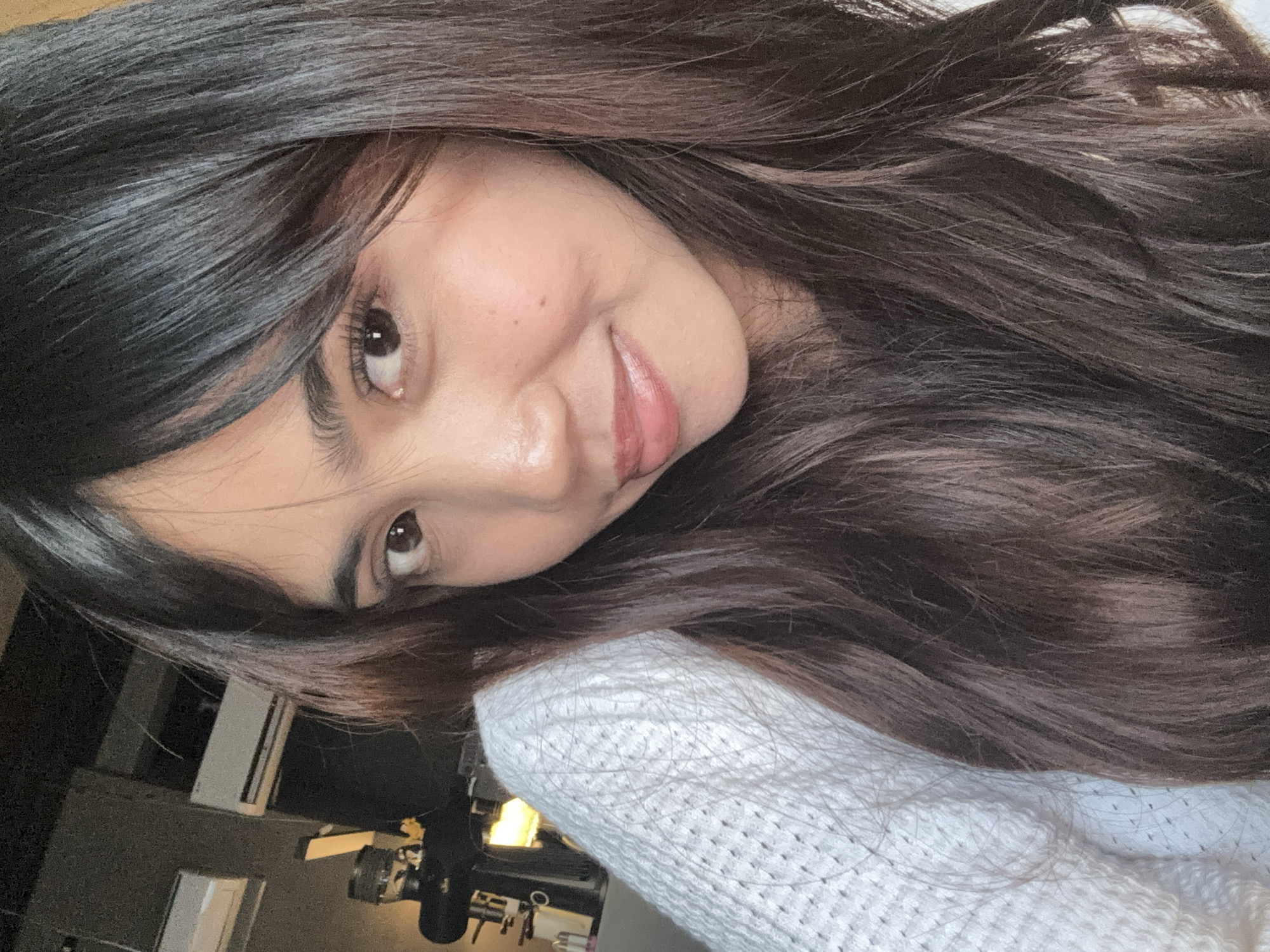 The image is a close-up selfie of a young woman with long, dark hair and fair skin. She is wearing a white, textured sweater. Her eyes are brown, and she has a slight smile on her lips. The background appears to be indoors, possibly a cafe or restaurant, with a dark wall and what looks like air conditioning units mounted high up. A dark object, possibly a coffee machine or dispenser, is visible on the left side of the frame. The lighting suggests an indoor environment, possibly with artificial light. The image does not provide any specific visual cues to confirm its location within Davao City, Philippines.