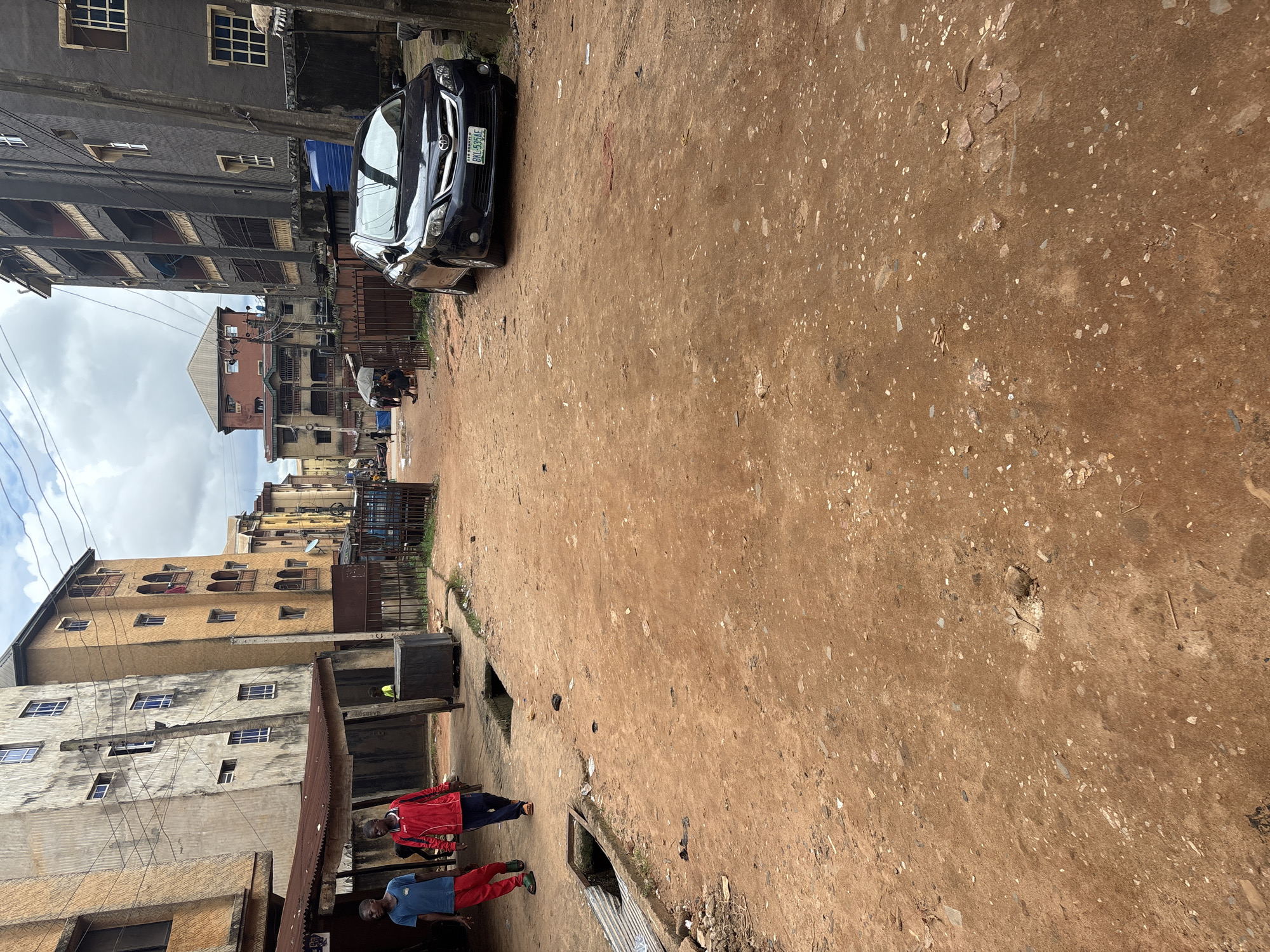 The image depicts a street scene in Onitsha, Nigeria, characterized by dusty, reddish-brown earth. Two young males are walking on the left side of the road. One, wearing a blue t-shirt and red pants, walks in front, while the other, in a red jacket and dark pants, follows behind. The surroundings show multi-story buildings with balconies and windows, typical of urban Nigerian architecture. A dark-colored sedan is parked on the right side of the road, its license plate visible. The scene suggests a typical day in a bustling urban environment, with elements of everyday life such as buildings, vehicles, and pedestrians. The presence of drainage ditches and some scattered debris indicates an unpaved and possibly underserved urban area.