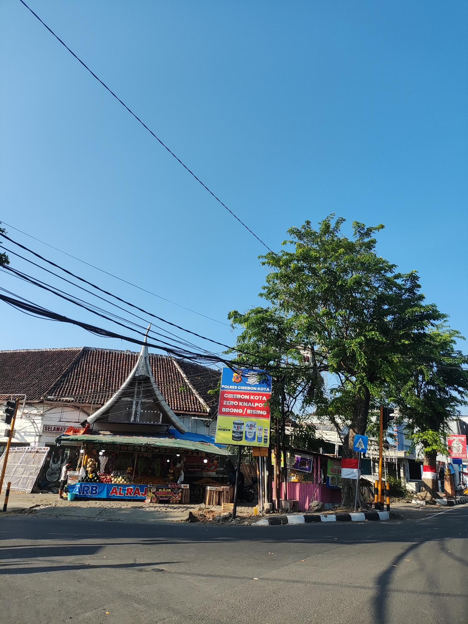 The image shows a street scene in Cirebon, Indonesia, on a clear day with a bright blue sky. In the foreground, a road is visible with tire tracks and shadows cast by trees. To the left, a fruit stand is set up under a blue awning, displaying a variety of fruits including bananas, apples, and what appear to be durians. The stand has signage that reads "ARB AL RAJ." Adjacent to the fruit stand is a building with a distinctive, ornate tiled roof. A traffic light is also present on the left side of the road.
Further to the right, a large, leafy green tree dominates the frame. Several power lines crisscross the sky above the scene. A tall, vertical sign with red and yellow lettering is visible behind the tree, advertising "POLRES CIREBON KOTA" and other details in Indonesian. A smaller blue sign with a white pedestrian symbol is mounted on a pole. In the background, more buildings and trees are seen, and there is a glimpse of what might be a motorcycle. The overall impression is a bustling, sunlit street in a residential or commercial area of the city.