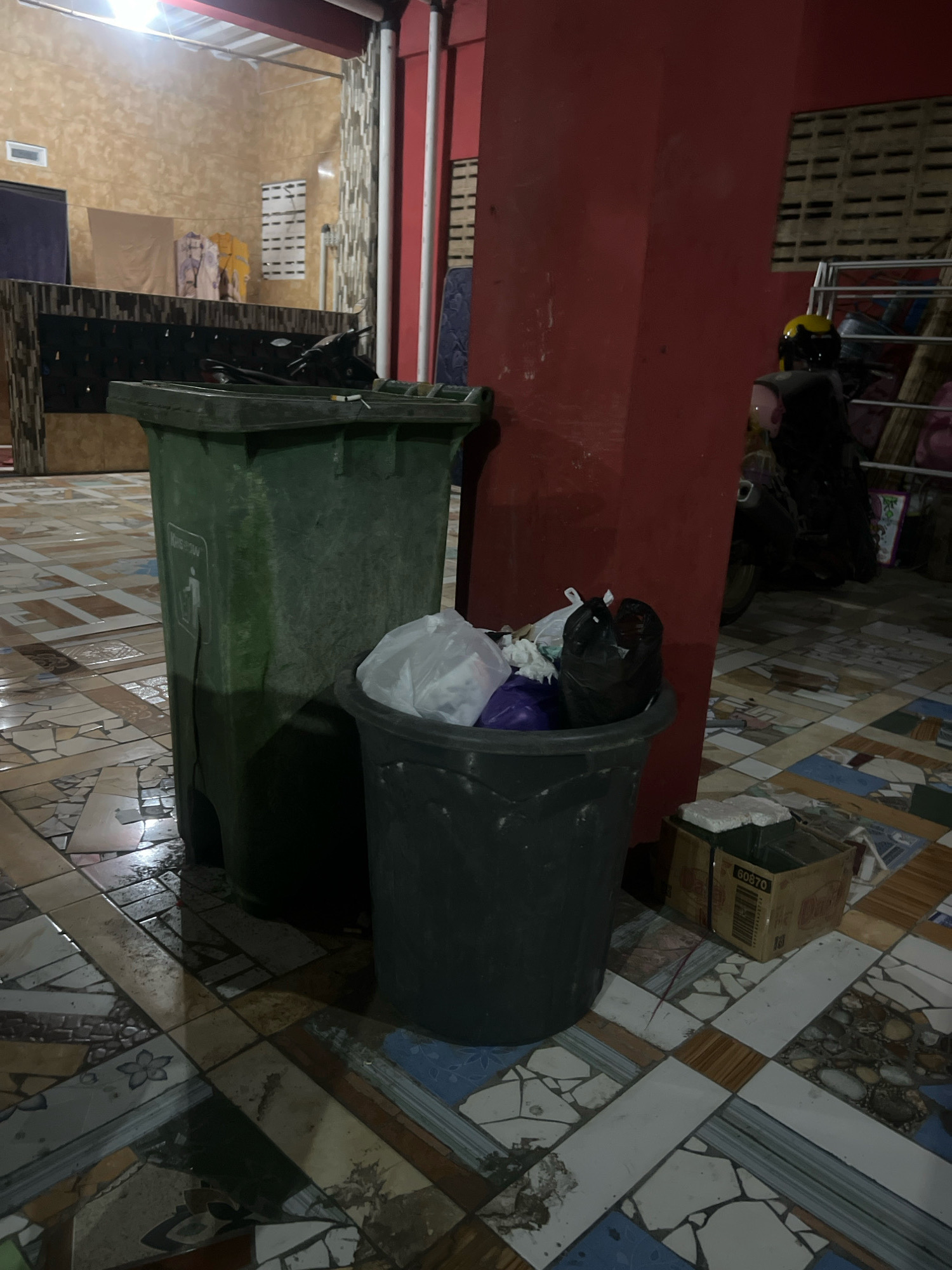 The image depicts an outdoor scene in Tlogoadi, Indonesia, likely a residential or commercial outdoor area, given the tiled flooring and building structure. In the foreground, two large green and gray trash bins are prominently placed. The green bin, positioned on the left, is taller and appears empty, while the gray bin to its right is overflowing with various bags of refuse, including black plastic bags and what appears to be white plastic sheeting.
Behind the trash bins, a red pillar supports an overhead structure, suggesting it is part of a building's facade or outdoor patio. To the right of the pillar, a motorcycle with a yellow helmet is partially visible, along with a drying rack holding laundry and other items, indicating domestic activity. In the background, a wall with a textured finish and some hanging garments can be seen. The floor is covered with decorative tiles, some of which appear wet or stained. A cardboard box labeled "60B70" is situated on the floor near the gray trash bin, also containing some white material. The overall impression is of a functional but somewhat cluttered outdoor space.