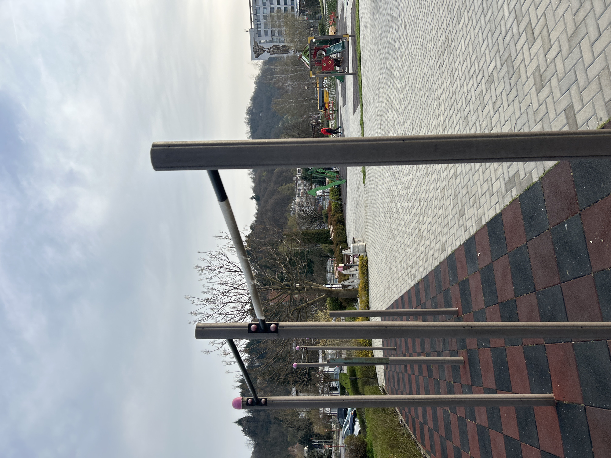 The image shows an outdoor workout area in Zlatograd, Bulgaria. Parallel bars of varying heights run along the right side of the frame, extending into the distance. The ground underneath is covered in alternating red and black square tiles, transitioning to gray brick pavers to the right. Beyond the bars is a green space and trees, with a background of low mountains and a cloudy sky. In the distance, a playground and buildings are visible. A person in a red jacket is visible in the playground area.