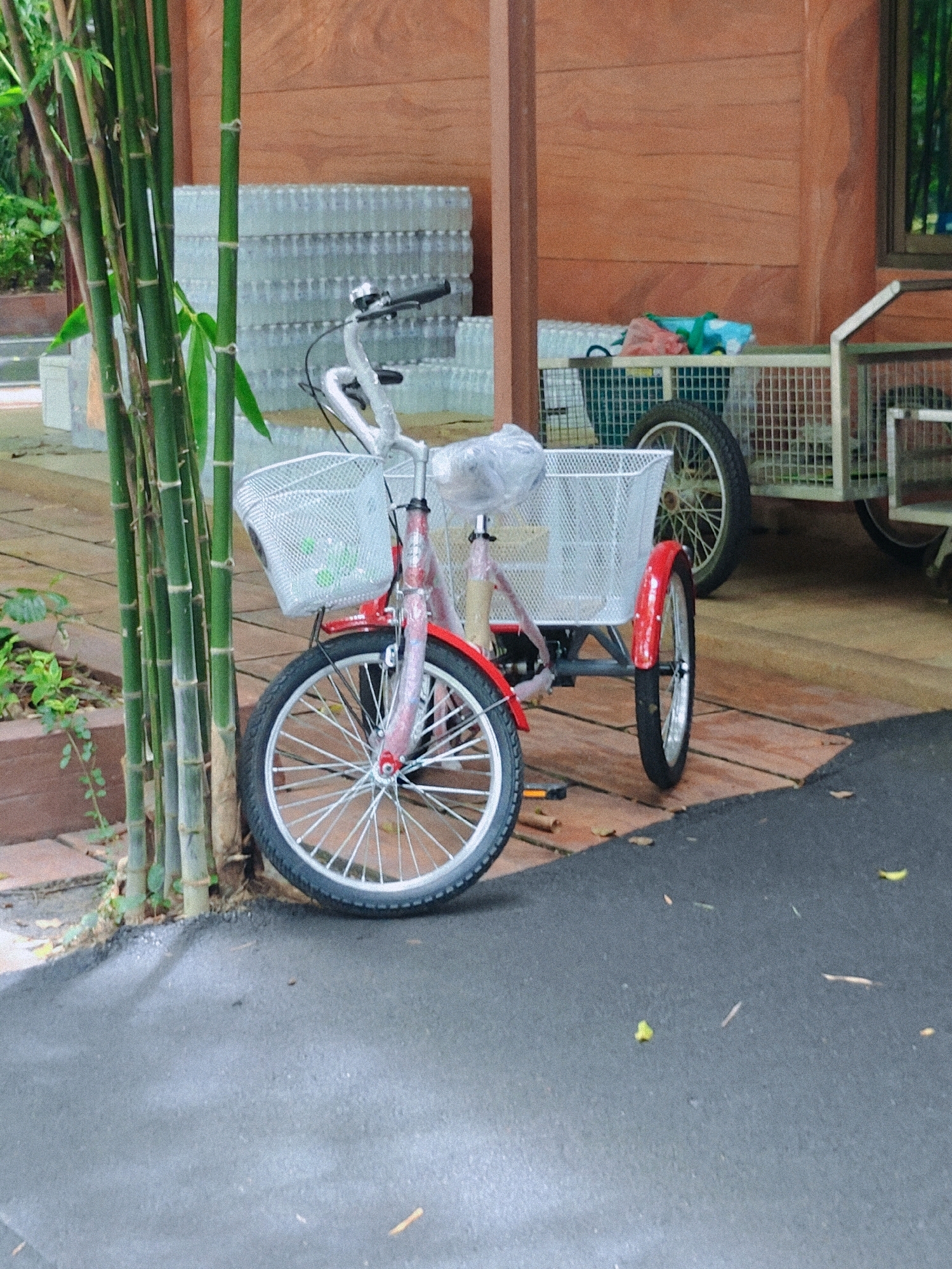 The image depicts a red and silver tricycle parked outdoors on a wooden deck adjacent to an asphalt path. The tricycle features two large baskets, one in the front and one in the rear, and is partially covered with plastic. To the left of the tricycle, a cluster of tall bamboo stalks stands upright. In the background, a structure with wooden paneling is visible, and stacked bottles of water are arranged against it. Further to the right, a wheeled cart with a wire mesh structure is also present. The scene appears to be in a public area, potentially a park or an establishment in Hantra Subdistrict, Thailand, given the presence of bamboo and the architectural style. No people are present in the image.