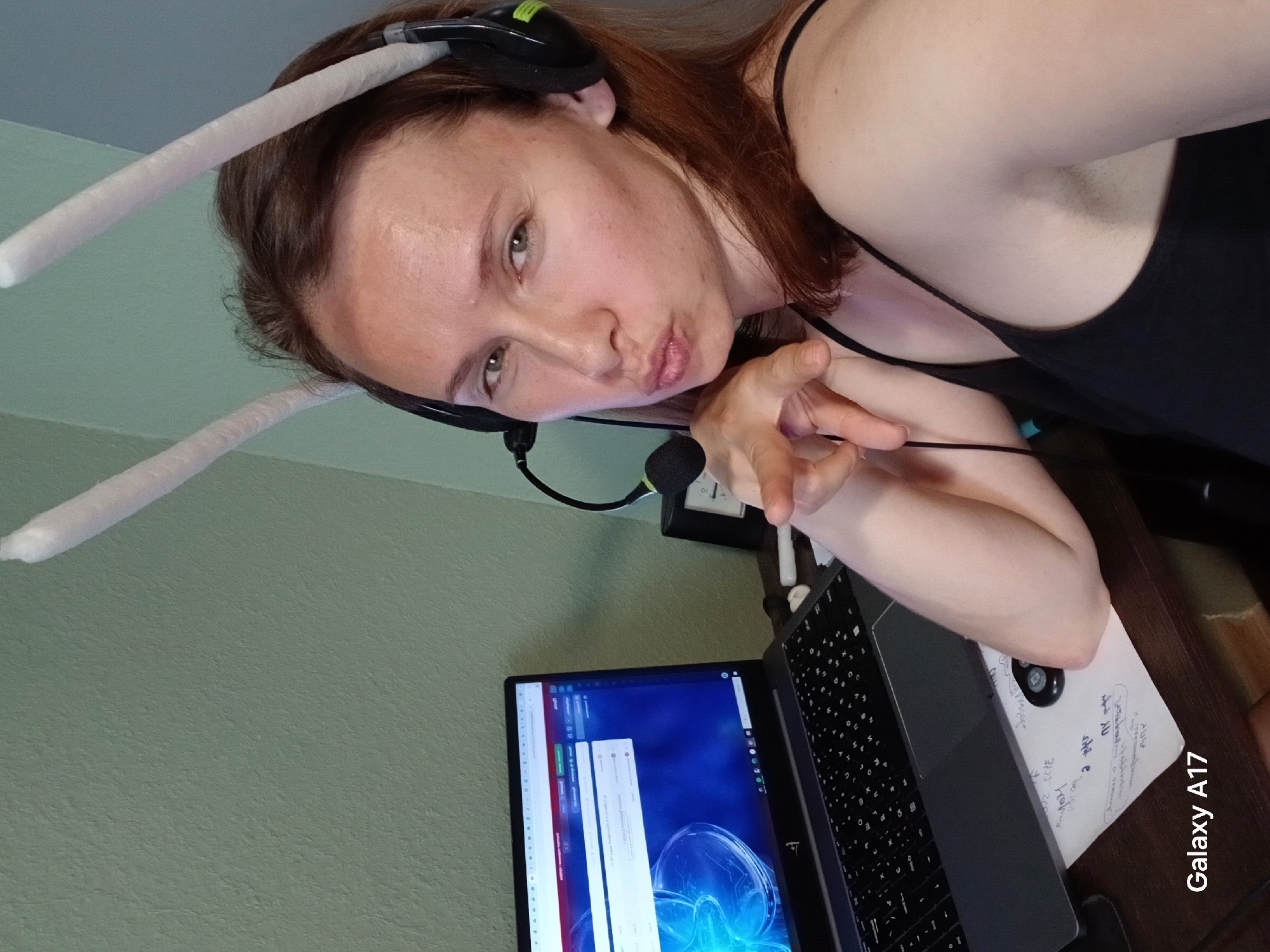 A woman with brown hair is wearing a black tank top and a headset with white padded antennae attached. She is making a kissy face and pointing at the camera. She is sitting at a desk with a laptop. A notebook labeled "Galaxy A17" is on the desk. The wall behind her is light green. Based on the "Galaxy A17" label and the Russian writing on the notebook, the location may be in Russia, but there is no evidence indicating it is specifically in Yekaterinburg.